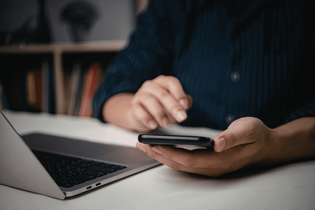 Person using a smartphone with a laptop nearby on a desk.