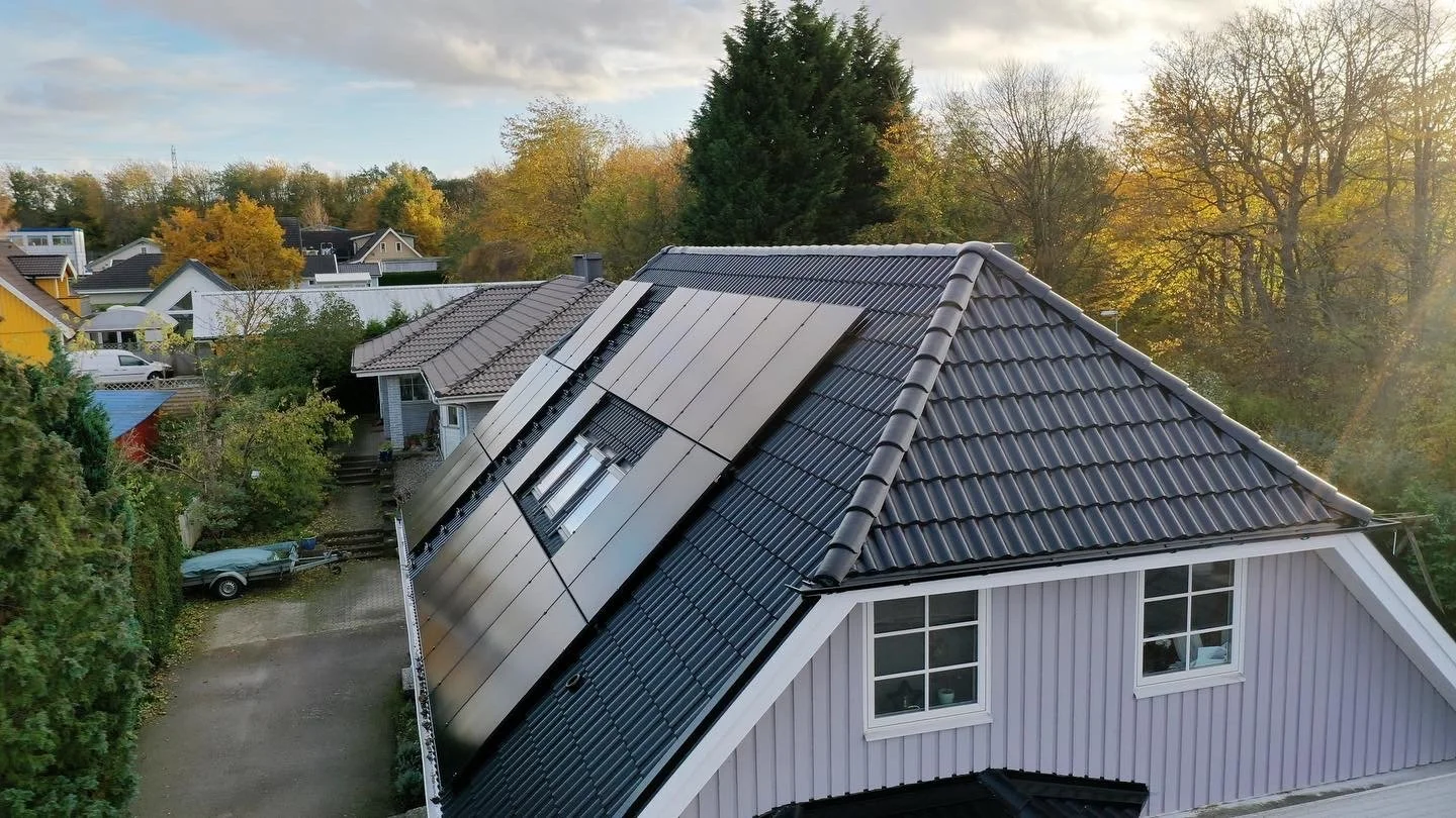 En takfönster är under installation på ett hus med två olika takmaterial, kopparfärgade takpannor och solpaneler, med en bakgrund av träd och andra hus i ett bostadsområde.