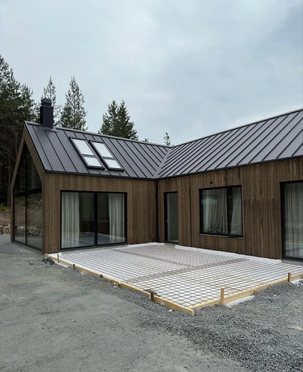 Nybyggt hus med träfasad, stora glasdörrar och fönster, samt ett metalltak. Området framför huset är under konstruktion, med en platt yta och stöd av träkanter.