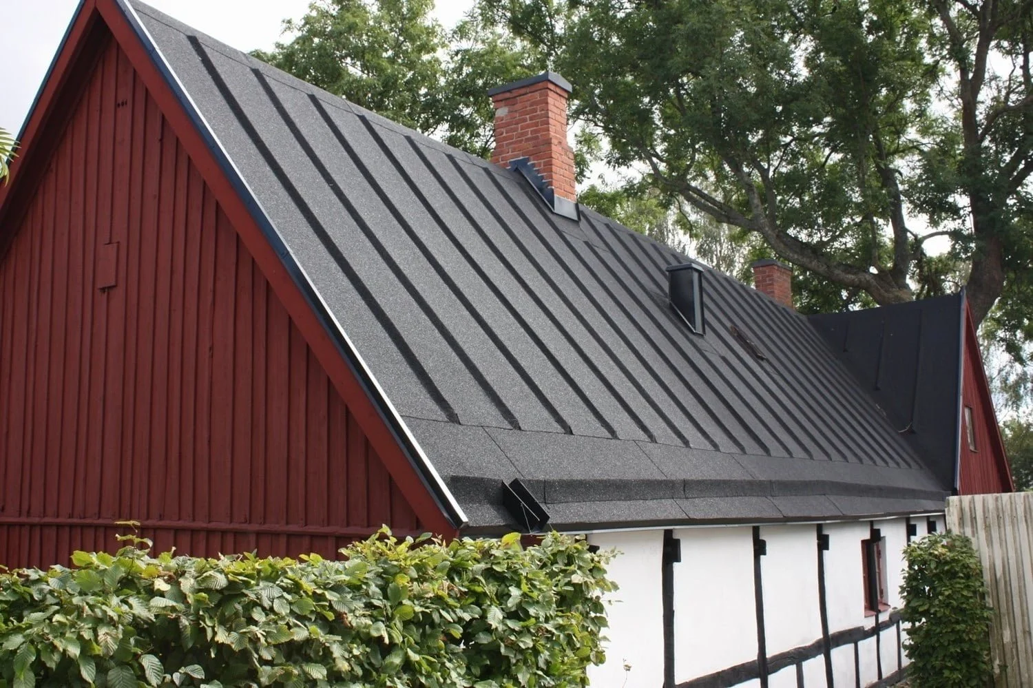 Hus med svart takbanna och röda väggar, omgiven av gröna träd och buskar.