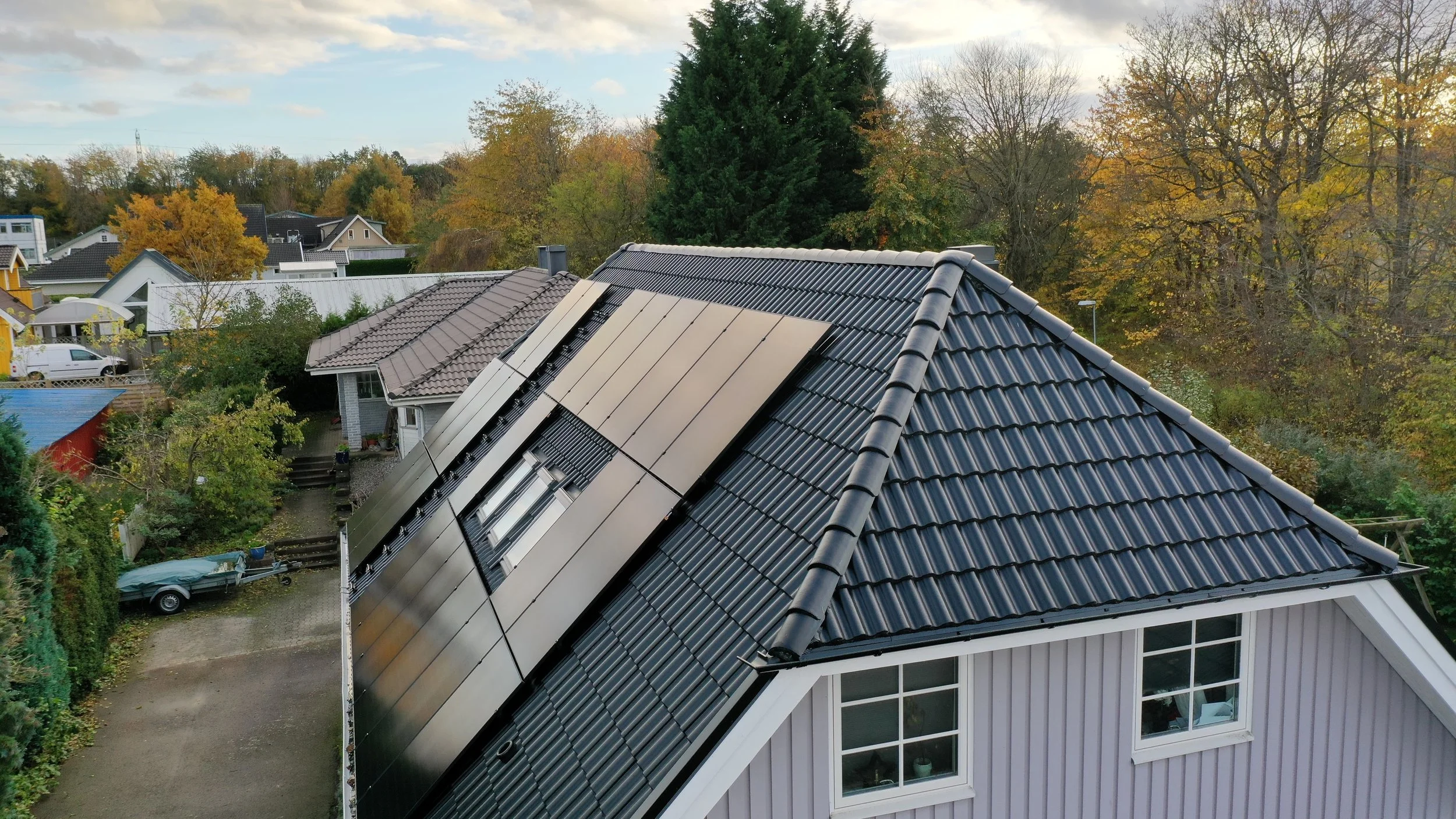 Husets tak med solpaneler och nya takpannor, omgiven av träd och grönska, i ett bostadsområde på hösten.
