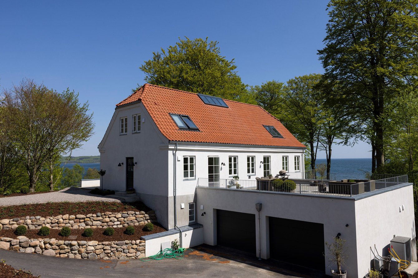 Vit modern villa med röd takterrass och utsikt över havet, omgiven av gröna träd.