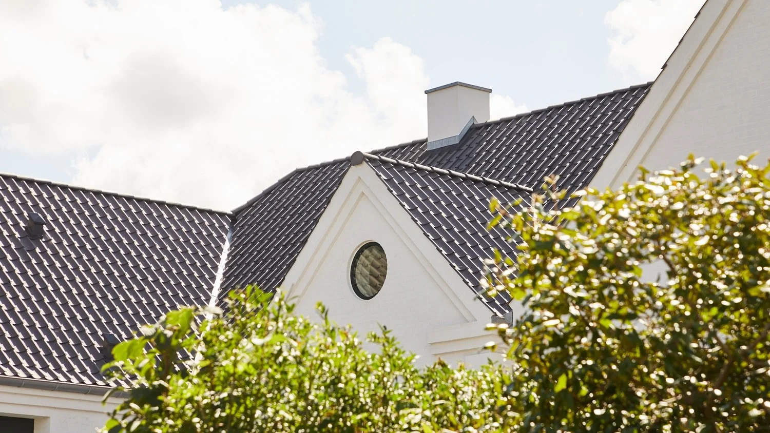 En vit villa med ett tak av mörkblått glaserat takpannor och en liten rund fönster. Det finns gröna buskar framför huset och himlen är blå med moln.