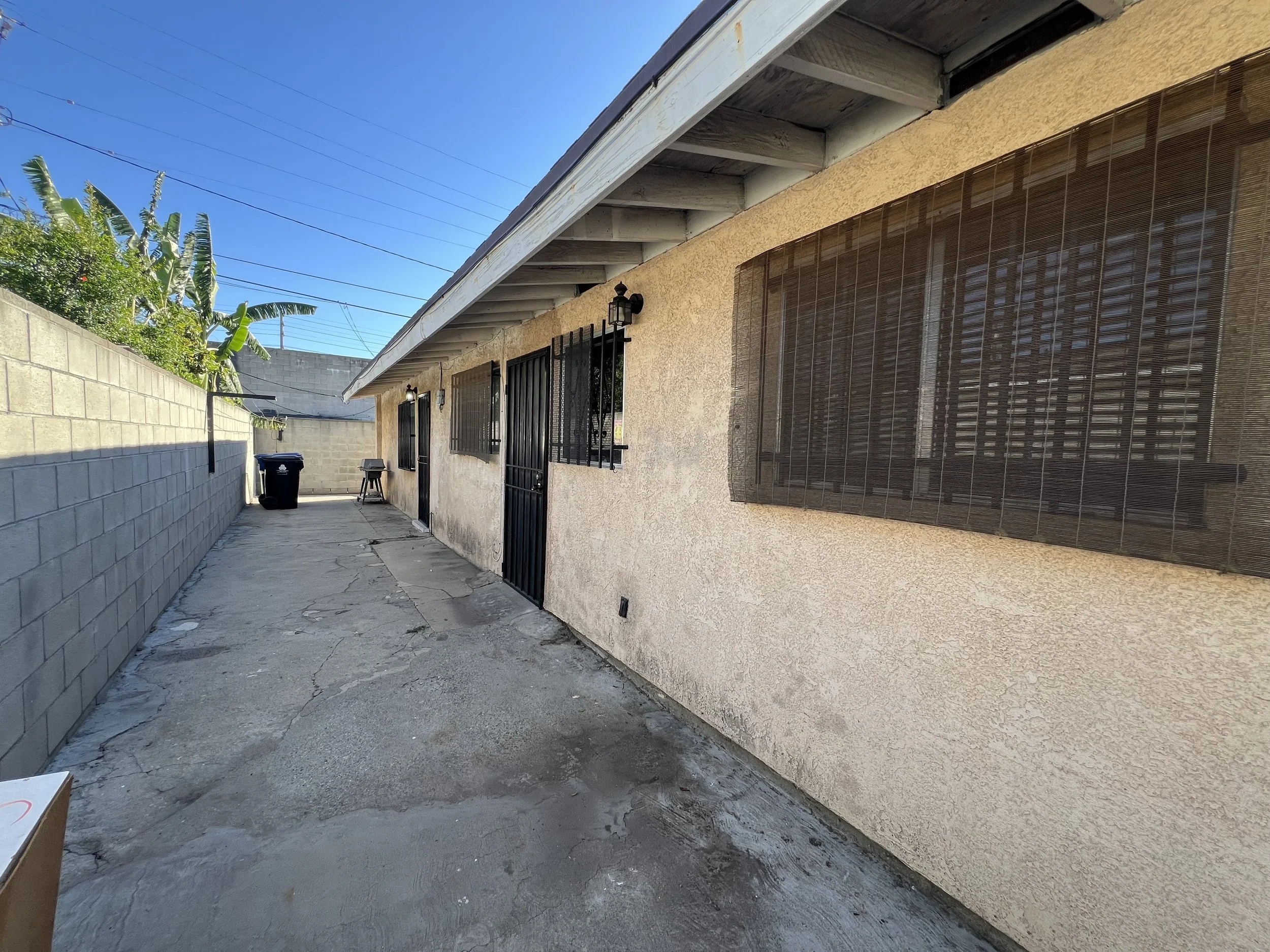 Backyard alleyway with a concrete ground, beige house wall with two windows and a door, barred on the windows, outdoor lanterns, and a bamboo shade on one window. A black trash bin and a small stool are also present. A concrete-block wall surrounds the alley with some green plants above it. The sky is clear and blue.