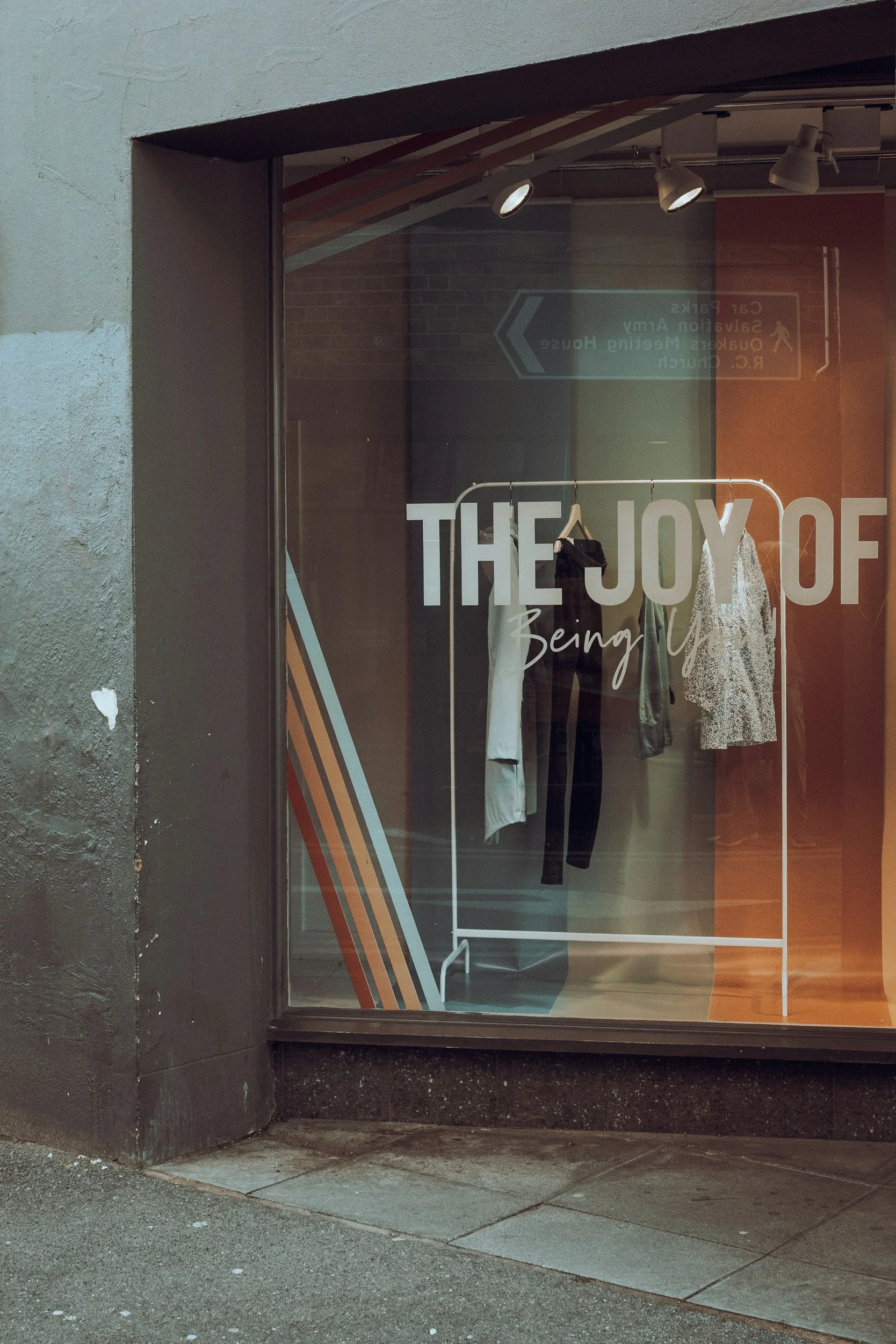 Store window displaying clothing on a rack, with painted text on the glass that reads 'THE JOY OF Being You.'