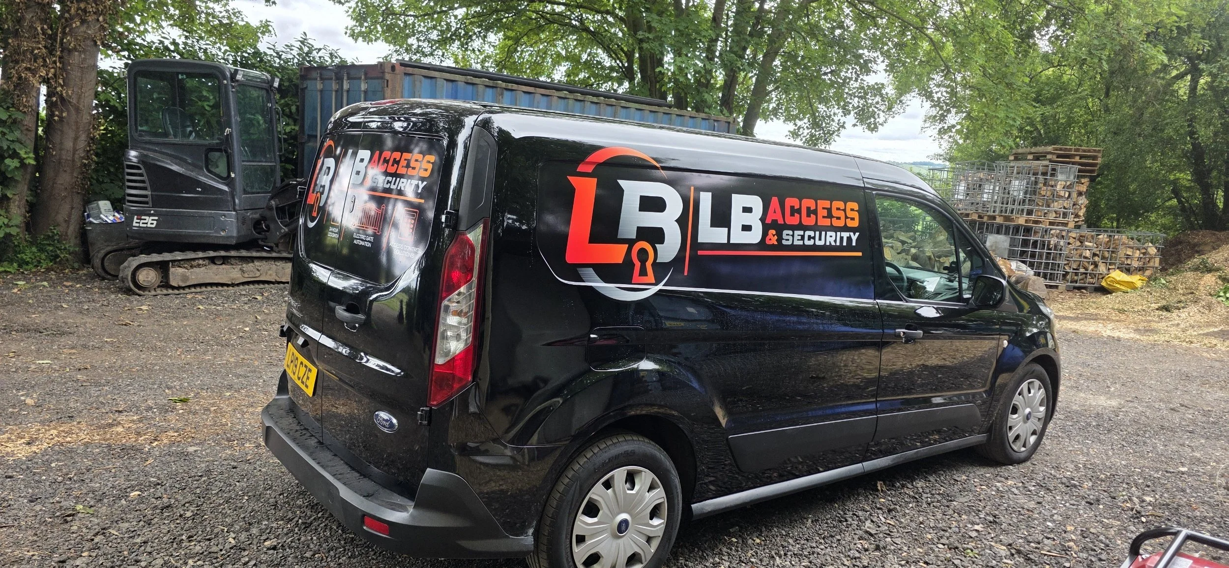 A black van and a small excavator parked on gravel with trees and stacked logs in the background. The van has the logo and name 'L3 LB Access & Security' on the side.