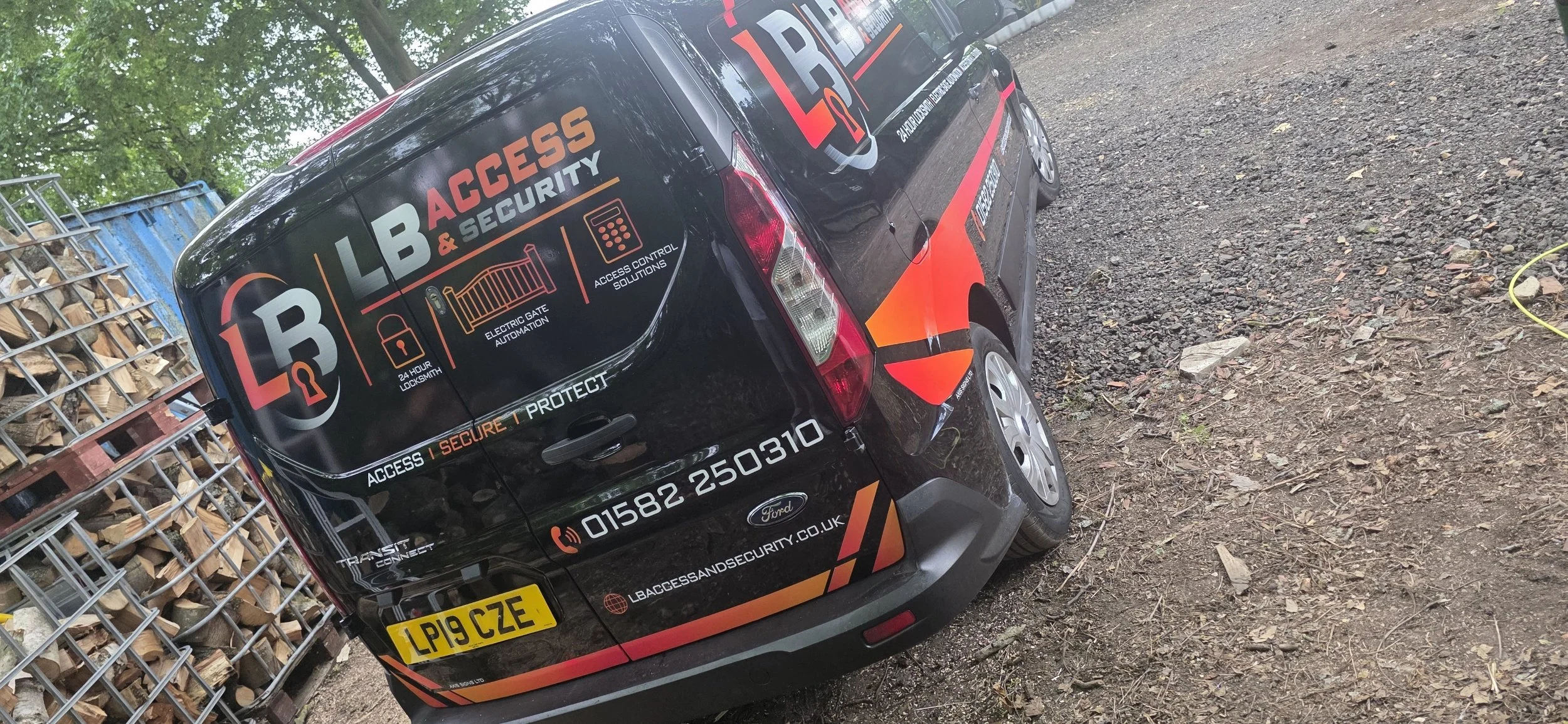 A black van with branding for LLB Access & Security, parked on a dirt surface beside a metal stacked firewood storage. The van displays services like 24-hour locksmith, electric gate automation, and access control solutions, along with a phone number and website.