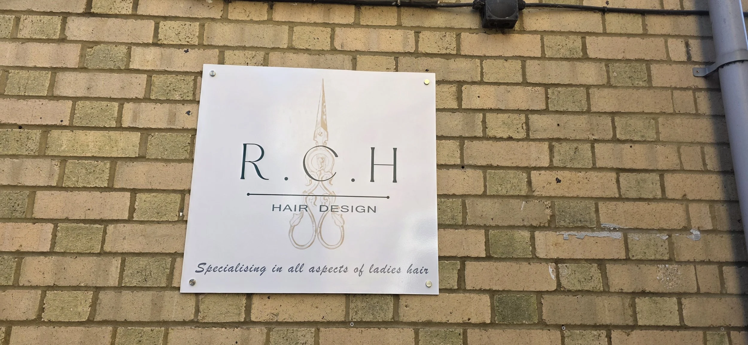 Sign for R. C. H Hair Design salon mounted on a brick wall, with text indicating it specializes in all aspects of ladies' hair.
