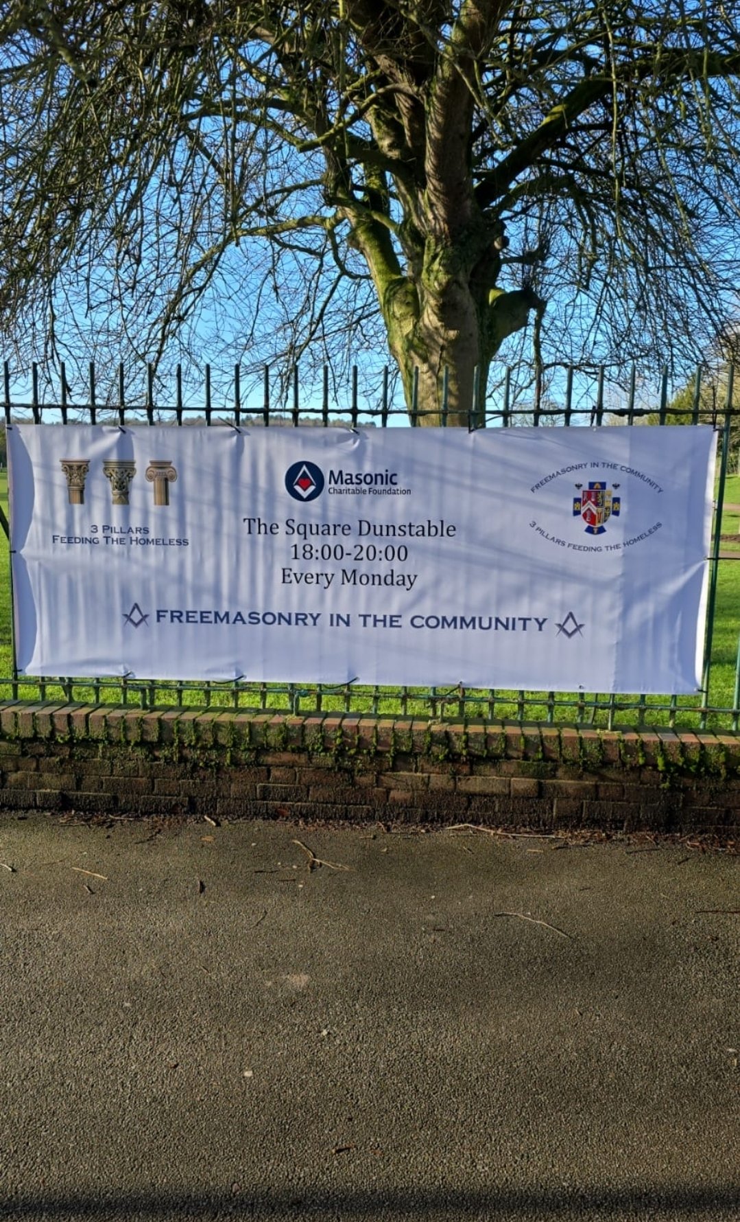 A large banner on a metal fence advertising a community event called 'The Square Dunstable,' organized by Masonry Charitable Foundation, happening every Monday from 6 pm to 8 pm at 3 Pillars Feeding The Homeless, with logos of Masonry and Freemasonry in the community, against a backdrop of a leafless tree and blue sky.