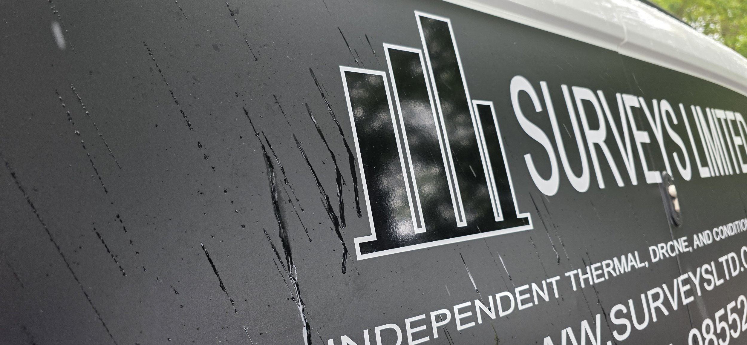 Close-up of a black waterproof sign with white text and a logo, showing water droplets on its surface.