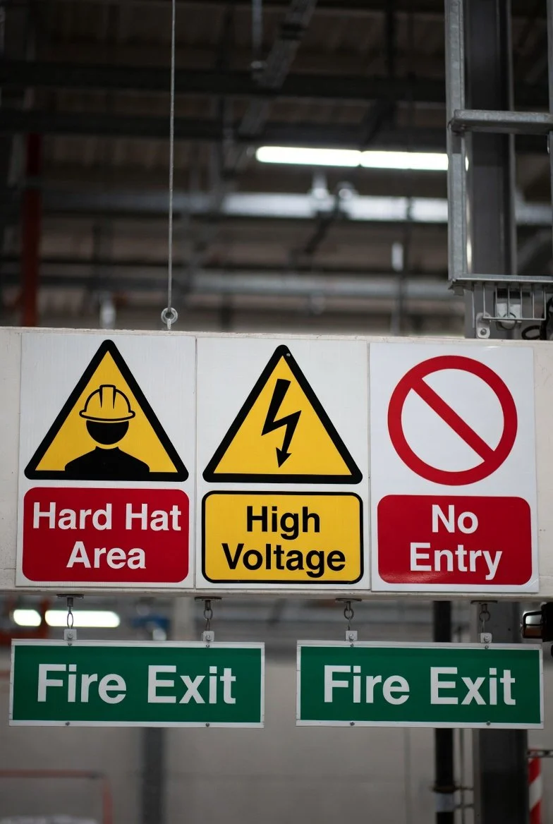 Warning signs in a factory or industrial area, indicating a hard hat area, high voltage, no entry, and fire exits.