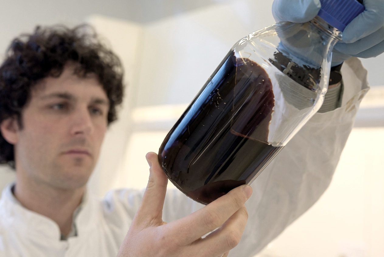 A scientist in a white lab coat and blue gloves examining a large glass jar filled with a liquified lignin.