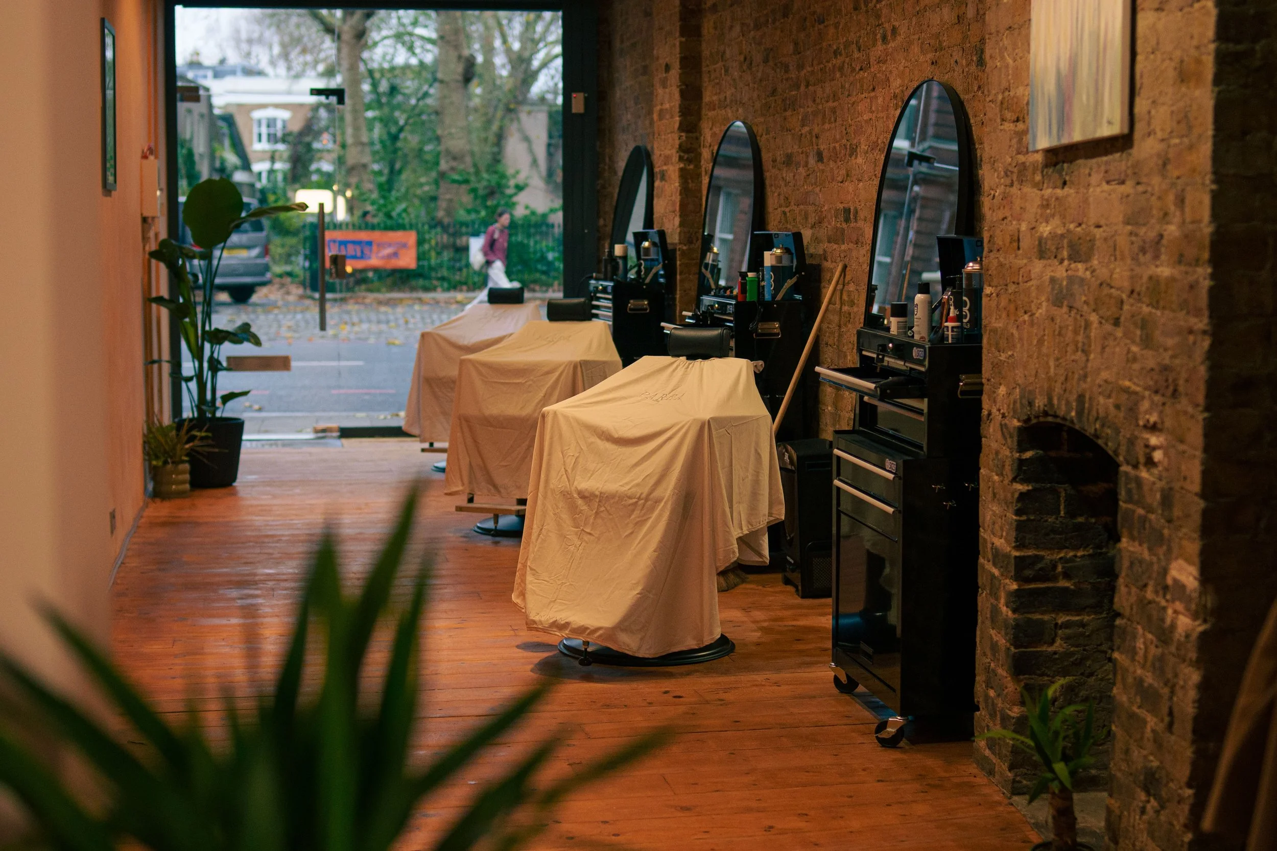 Interior of a hair salon with three styling stations covered with beige cloths, large mirrors on a brick wall, and a wooden floor, with a view of the street outside through the front window.