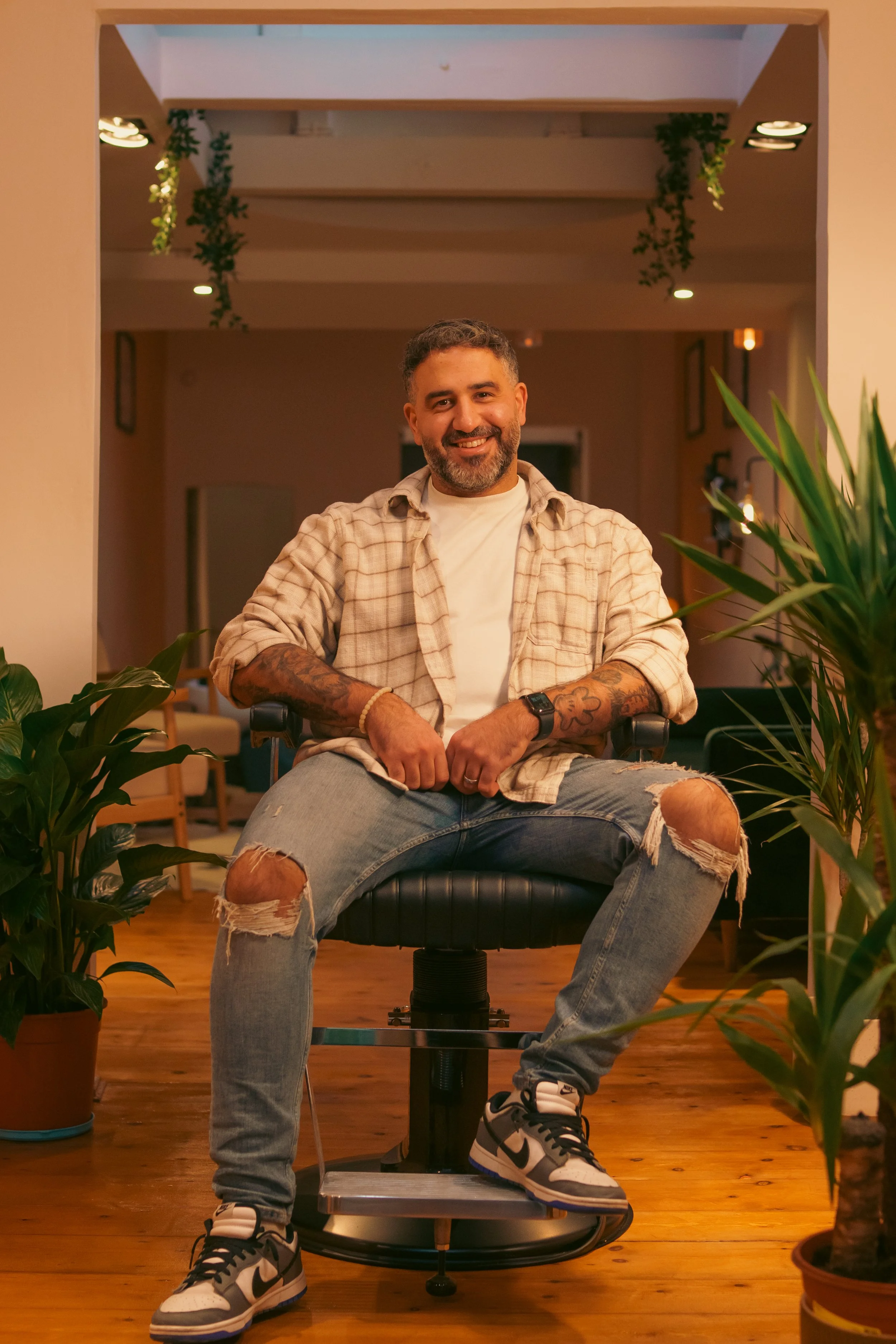 A smiling man with tattoos on his arms, wearing a beige plaid shirt, white t-shirt, ripped jeans, and sneakers, sitting in a chair in a cozy room surrounded by plants.