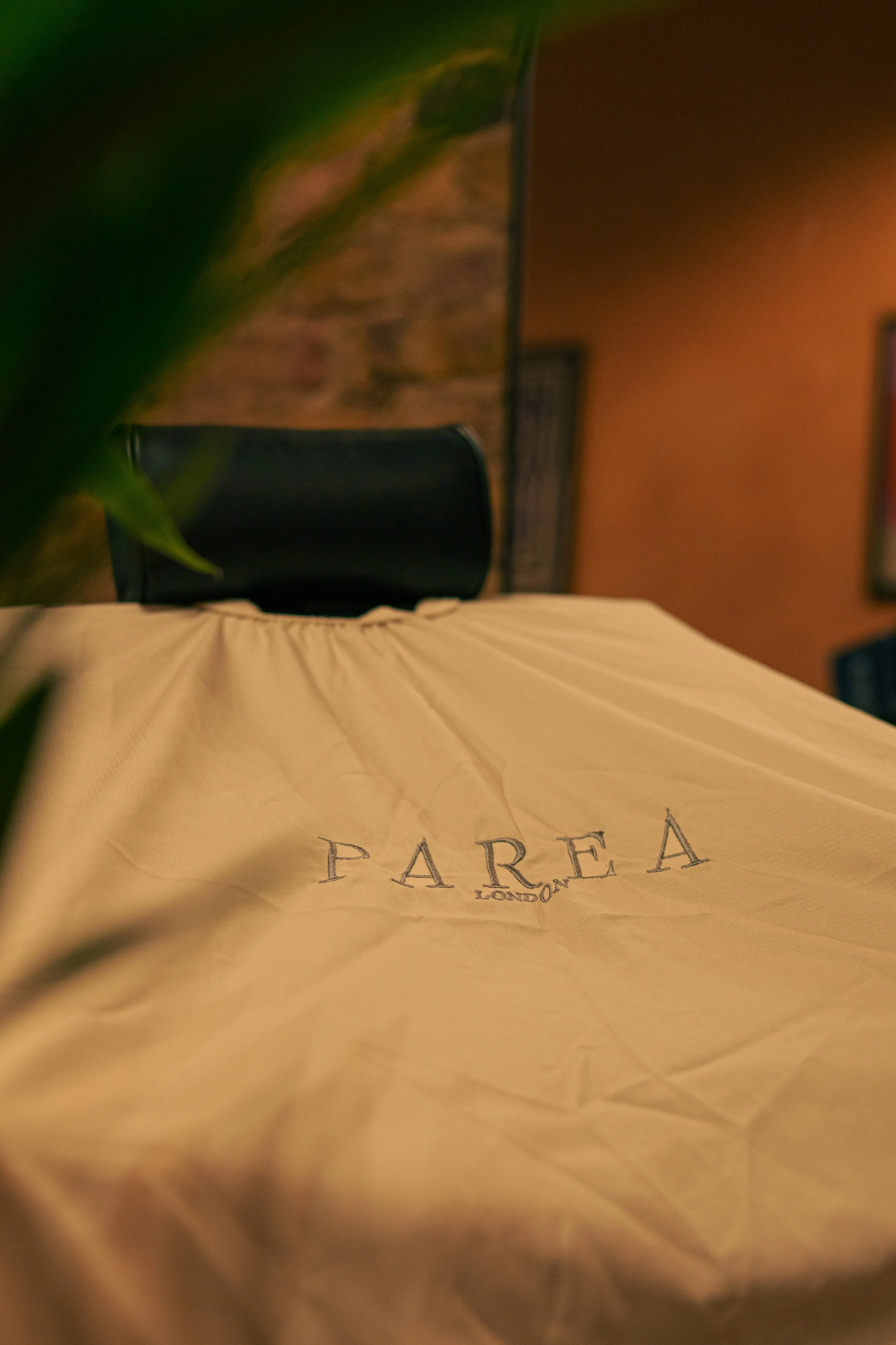 A beige tablecloth with the word 'PAREA' and 'LONDON' embroidered on it in front of a black chair, with a blurred background.