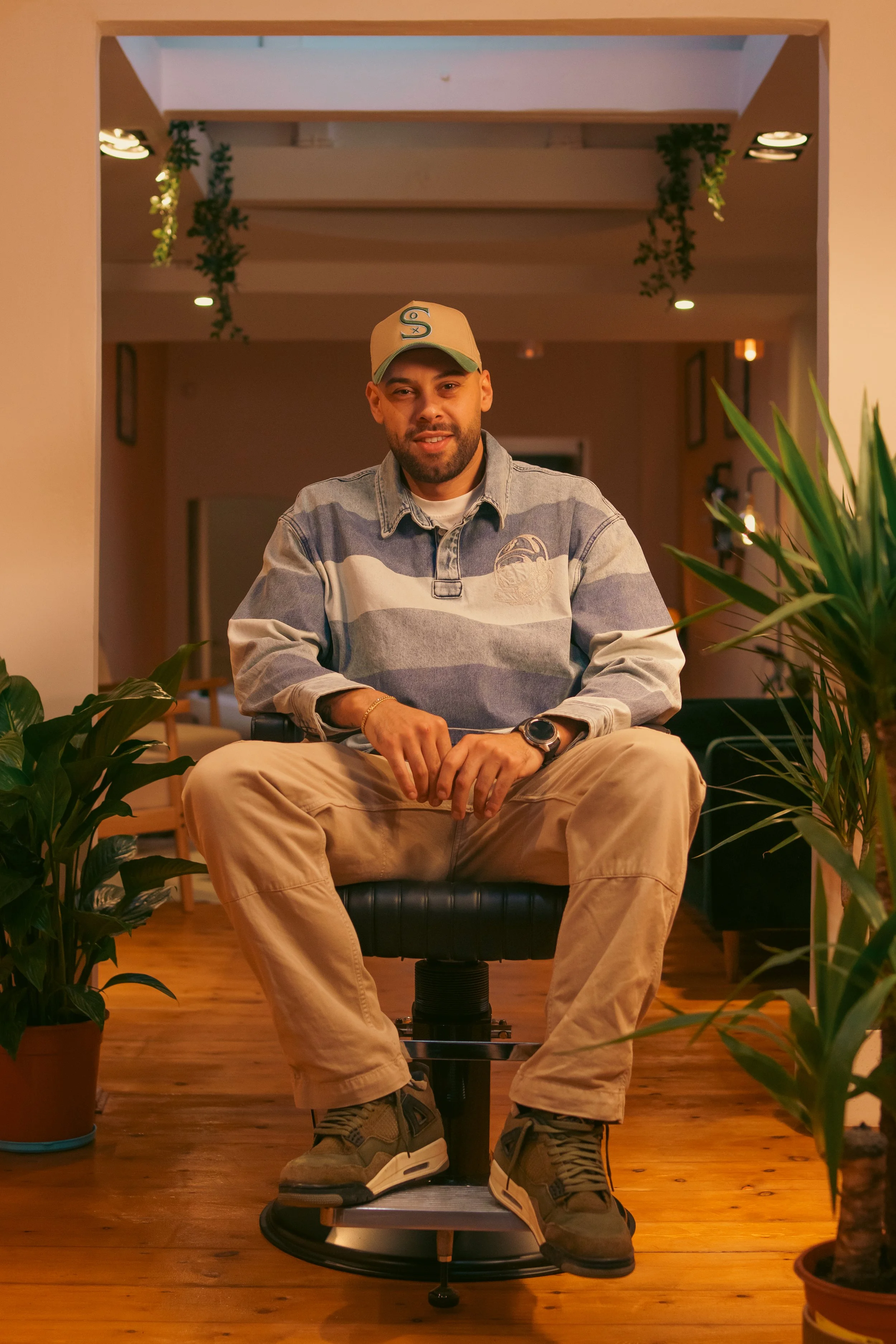 A man sitting on a black modern chair in a cozy indoor setting with plants on either side, warm lighting, and visible wooden flooring, wearing casual clothing including a baseball cap, striped shirt, beige pants, and sneakers.