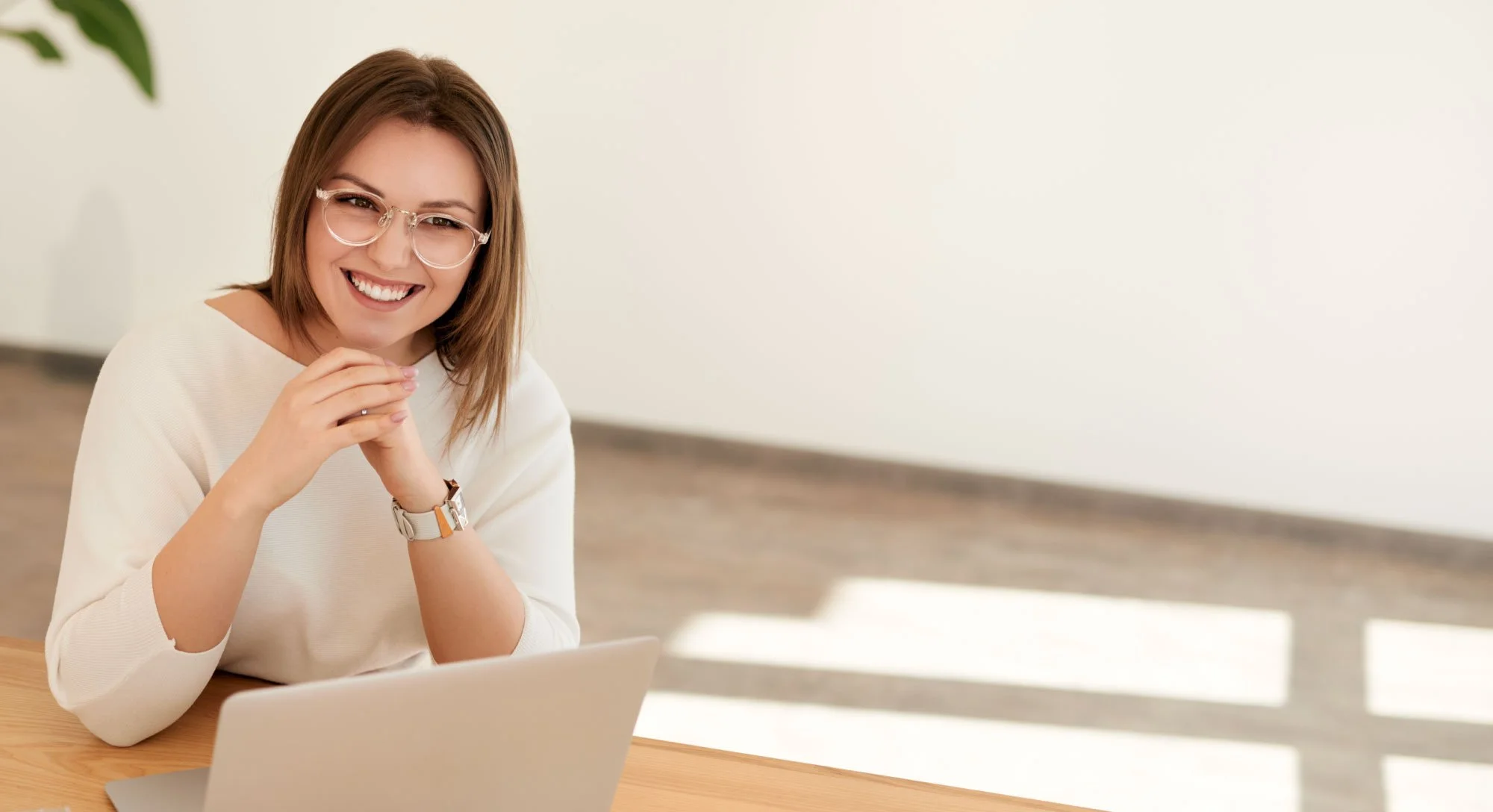 A smiling woman with glasses and a watch sitting at a desk with a laptop, in a well-lit room.