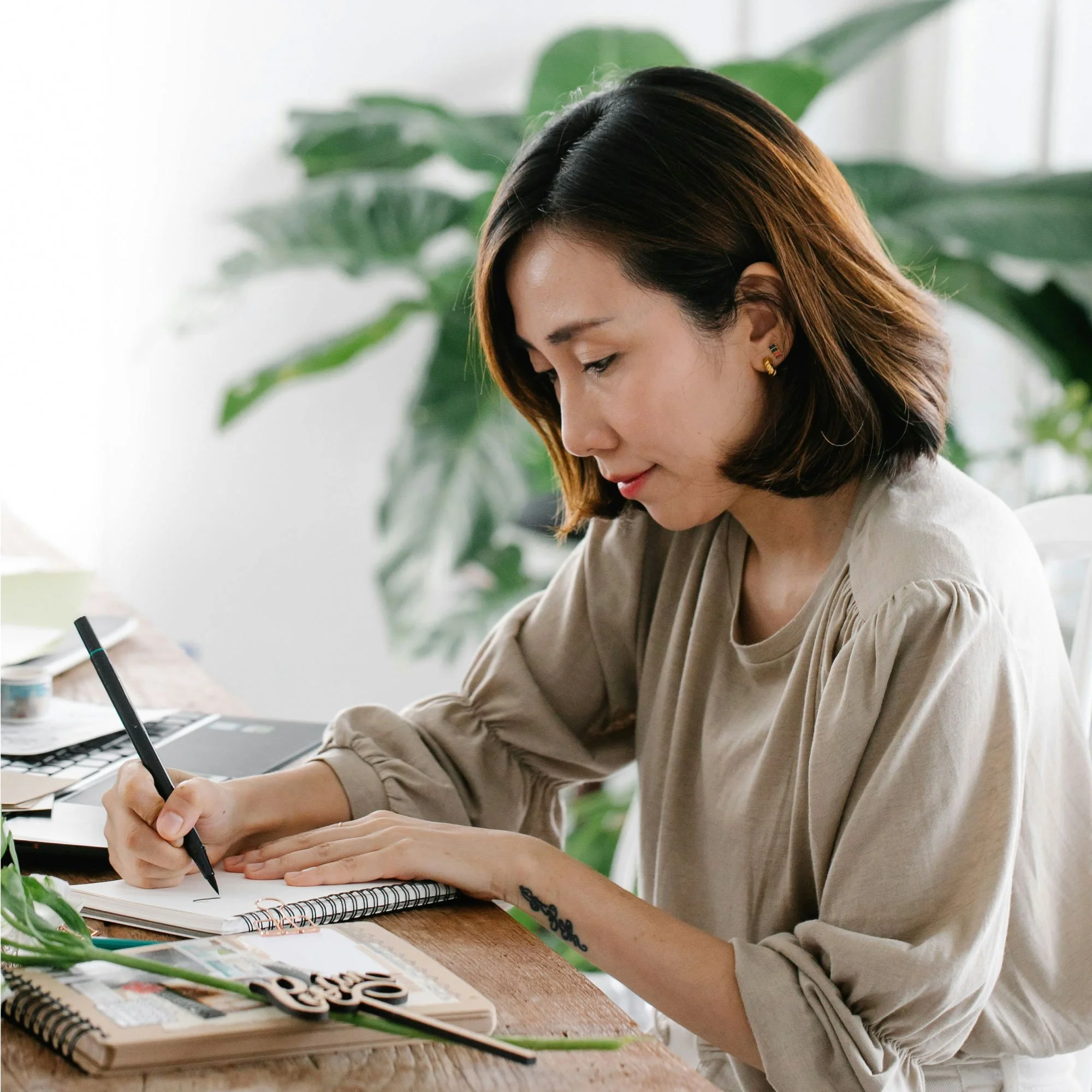 A woman writing in a notebook at a wooden table, with plants in the background.