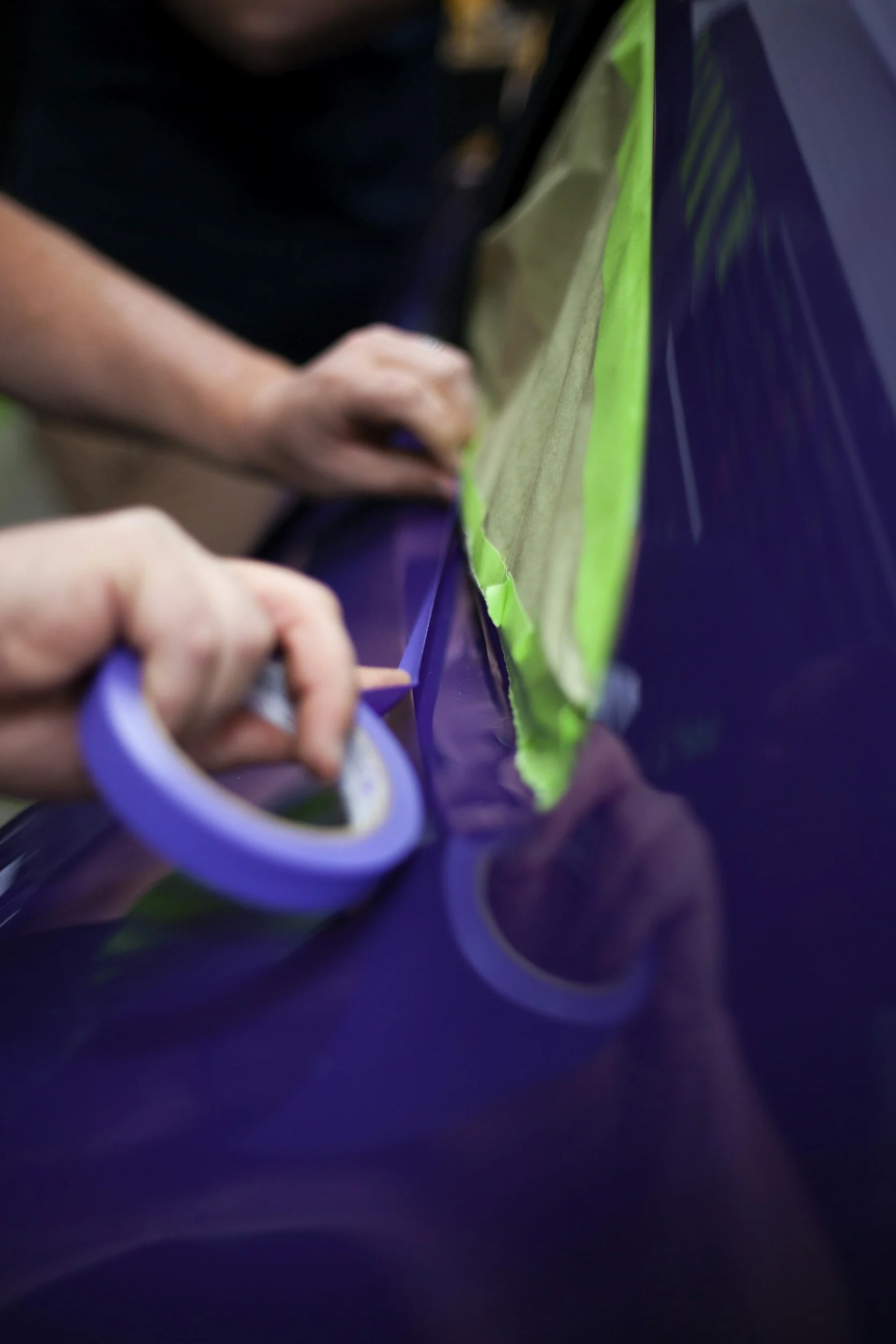 A person applying purple masking tape along the edge of a vehicle's purple painted surface, with green painter's tape and paper taped nearby.