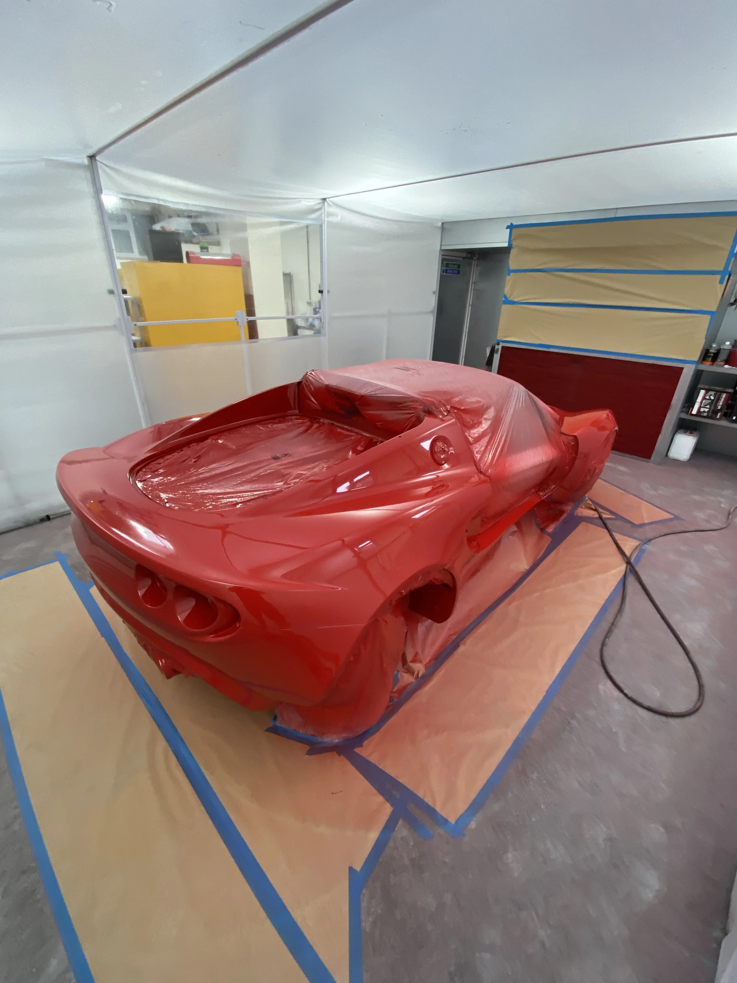 A red sports car is covered with plastic in a spray booth, ready for painting or drying.