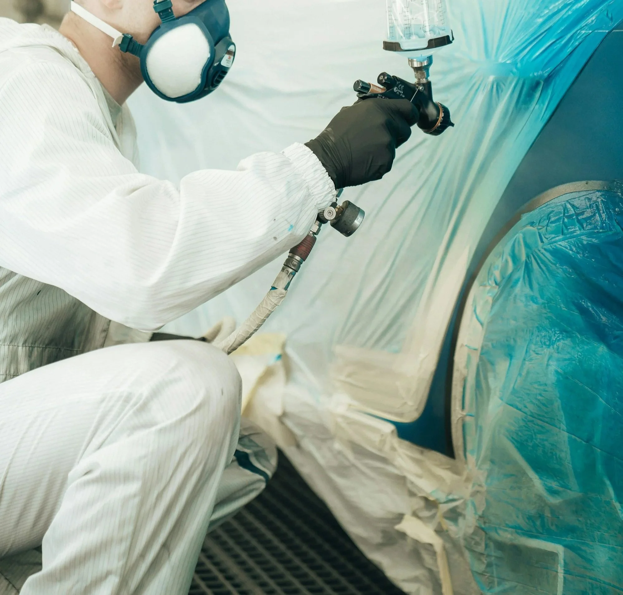 A person wearing protective gear, including a mask, gloves, and coveralls, spray-painting or coating a surface in a controlled environment.