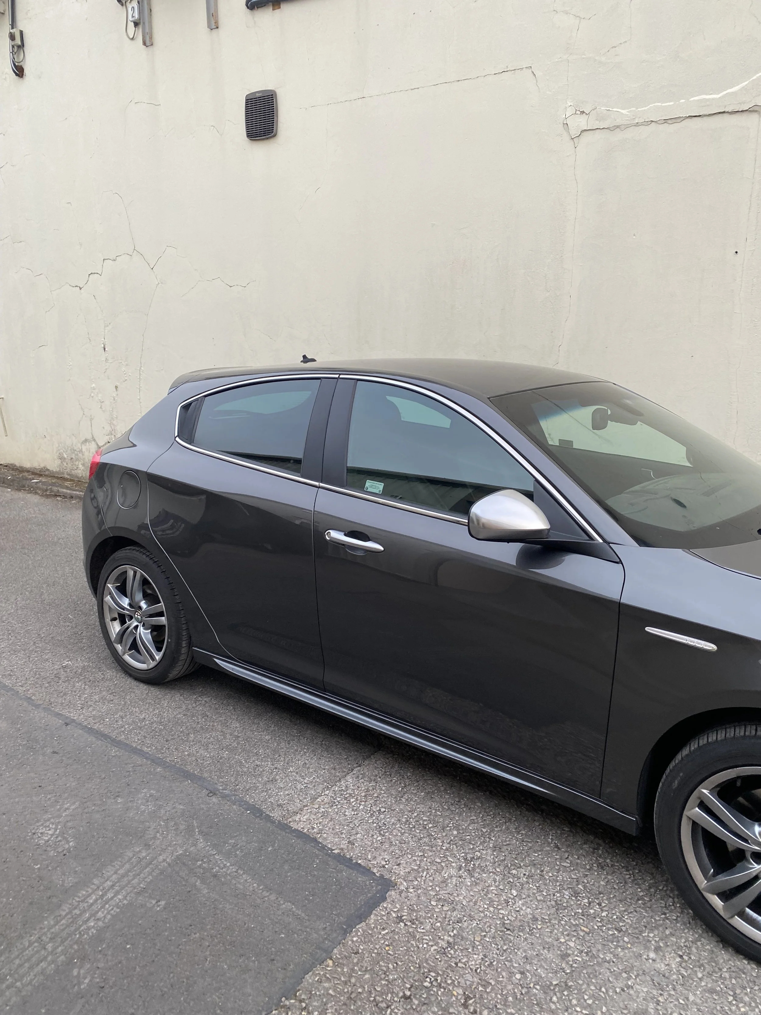 Black compact hatchback car parked on the street next to a beige wall with cracks.