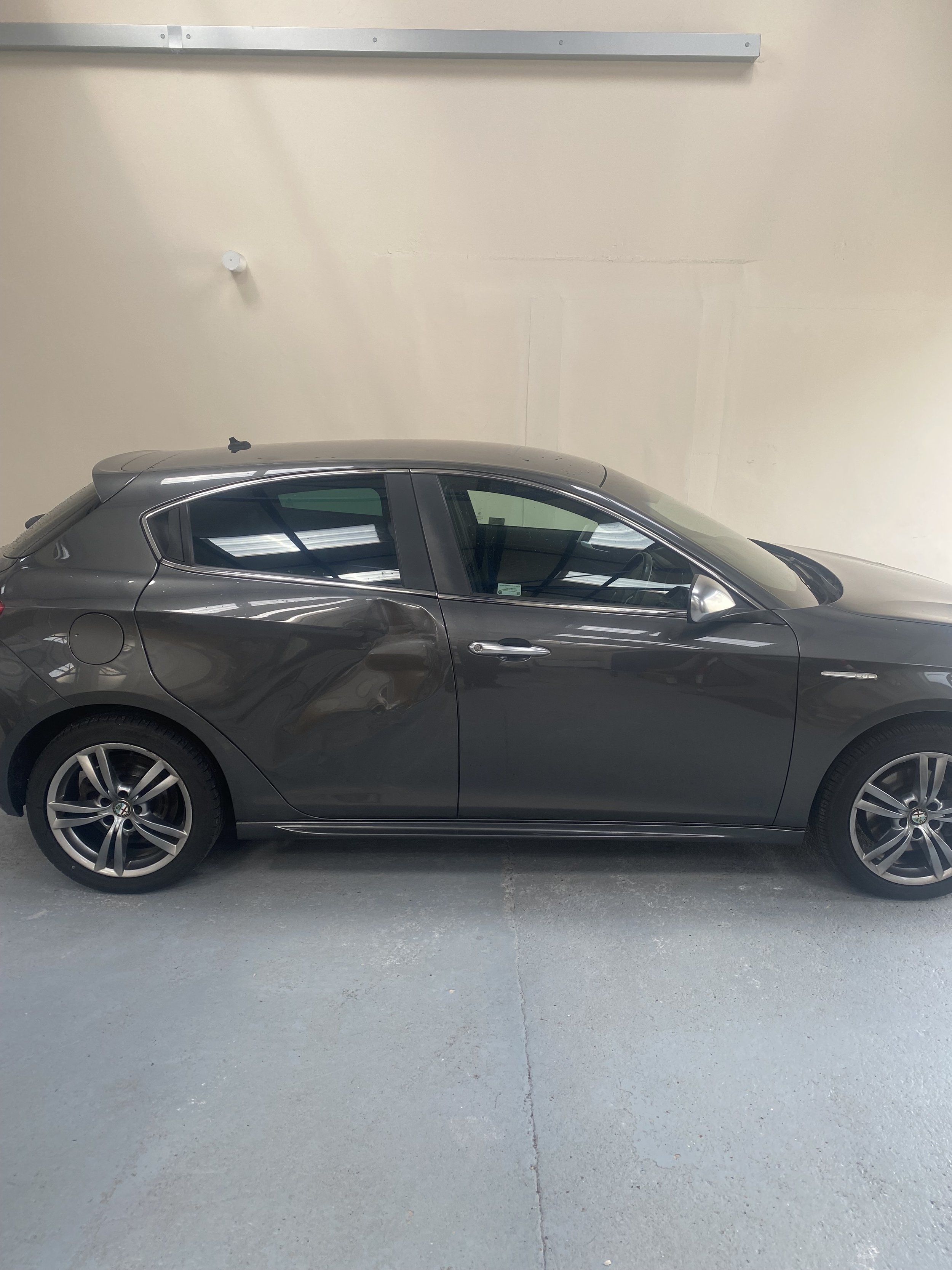 A black or dark gray compact hatchback car parked indoors against a beige wall with some garage or warehouse features.
