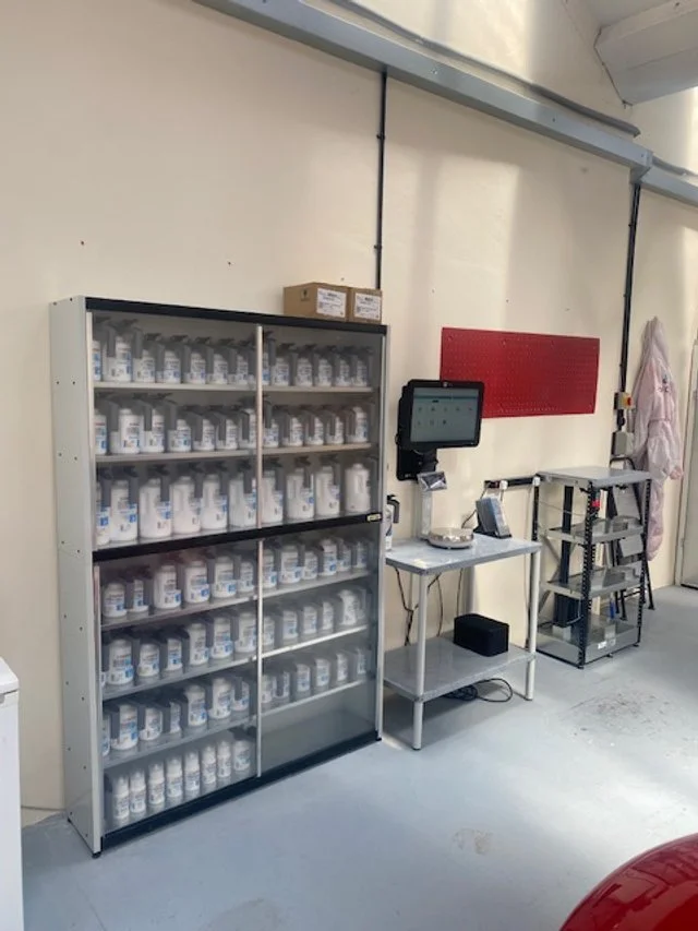 A room with a cabinet full of bottles, a computer monitor, a small table, and a metal rack, with a red panel on the wall and a hospital gown hanging on the wall.