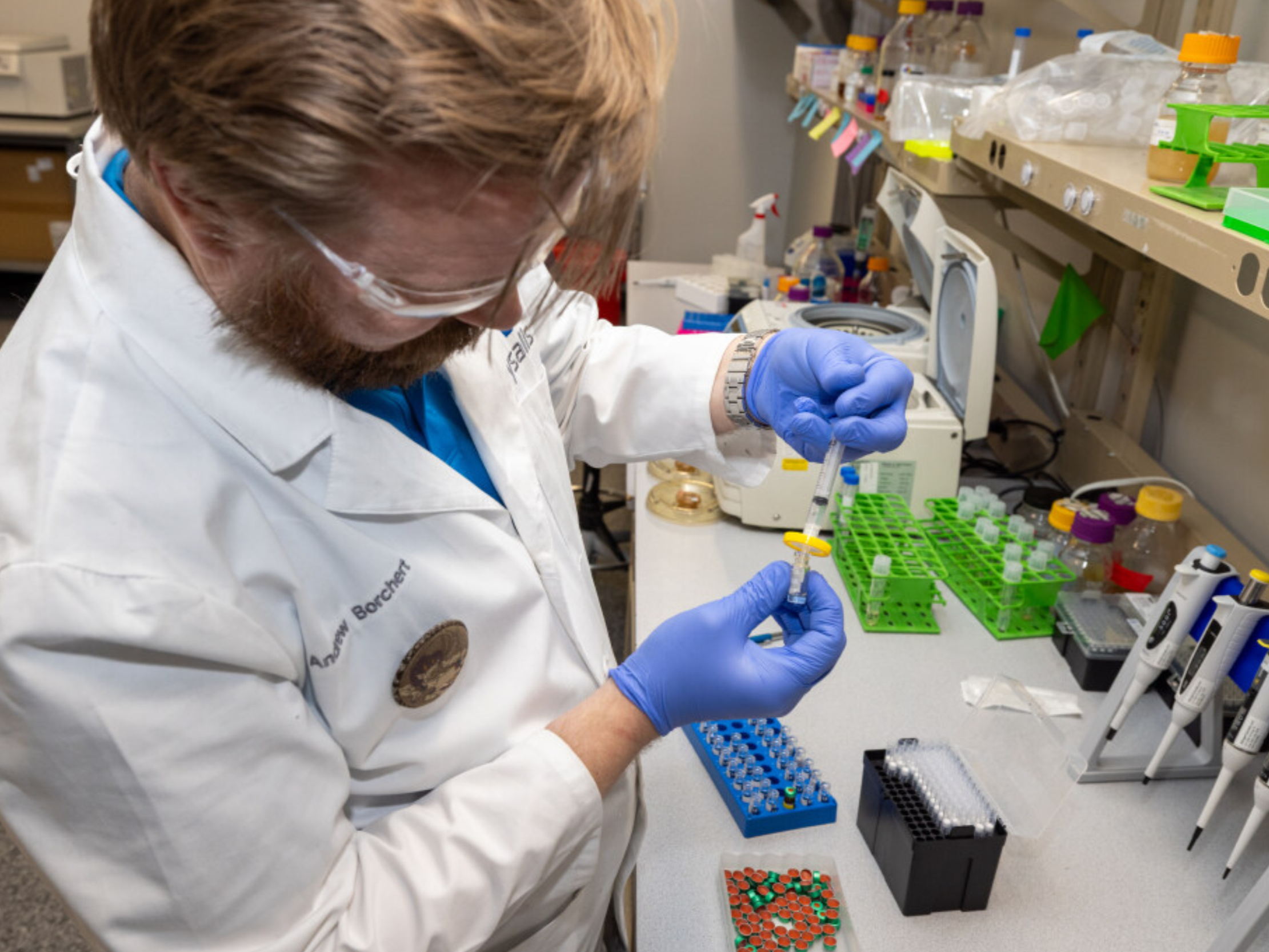 A scientist in a lab coat and blue gloves using a pipette to transfer liquid into a small test tube, surrounded by laboratory equipment and supplies.