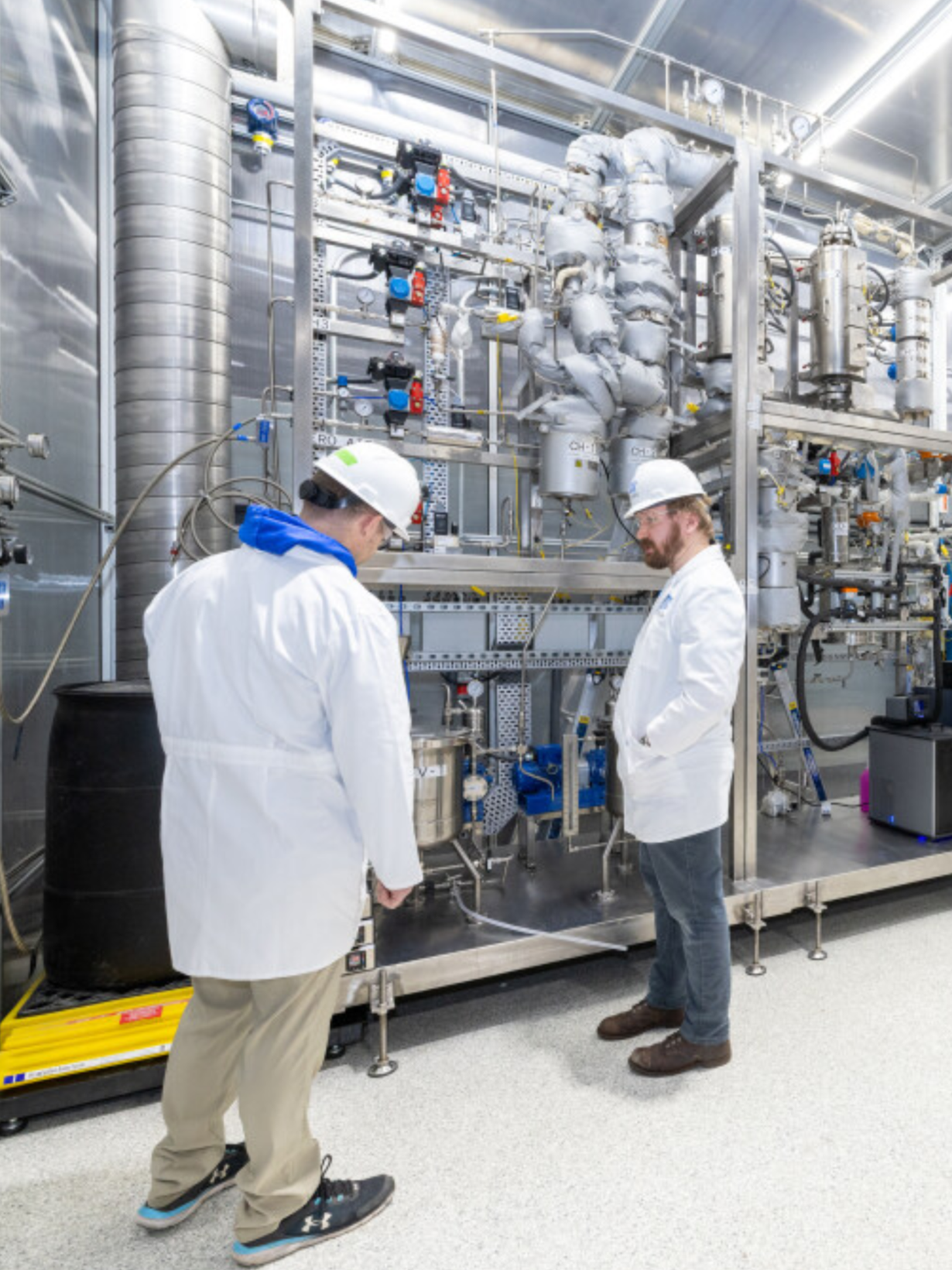 Two engineers or scientists in white lab coats and hard hats working in a high-tech industrial facility with complex machinery and piping.