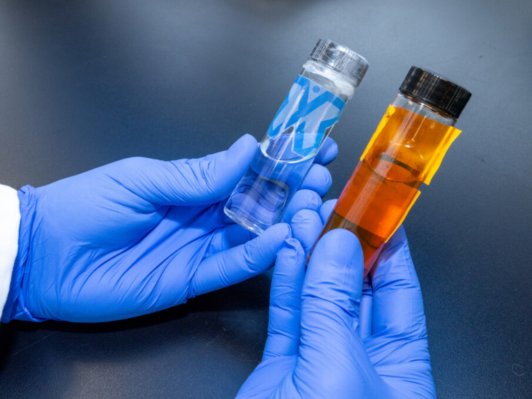 Person wearing blue gloves holding a small clear container with a blue and white label and a test tube filled with orange liquid.
