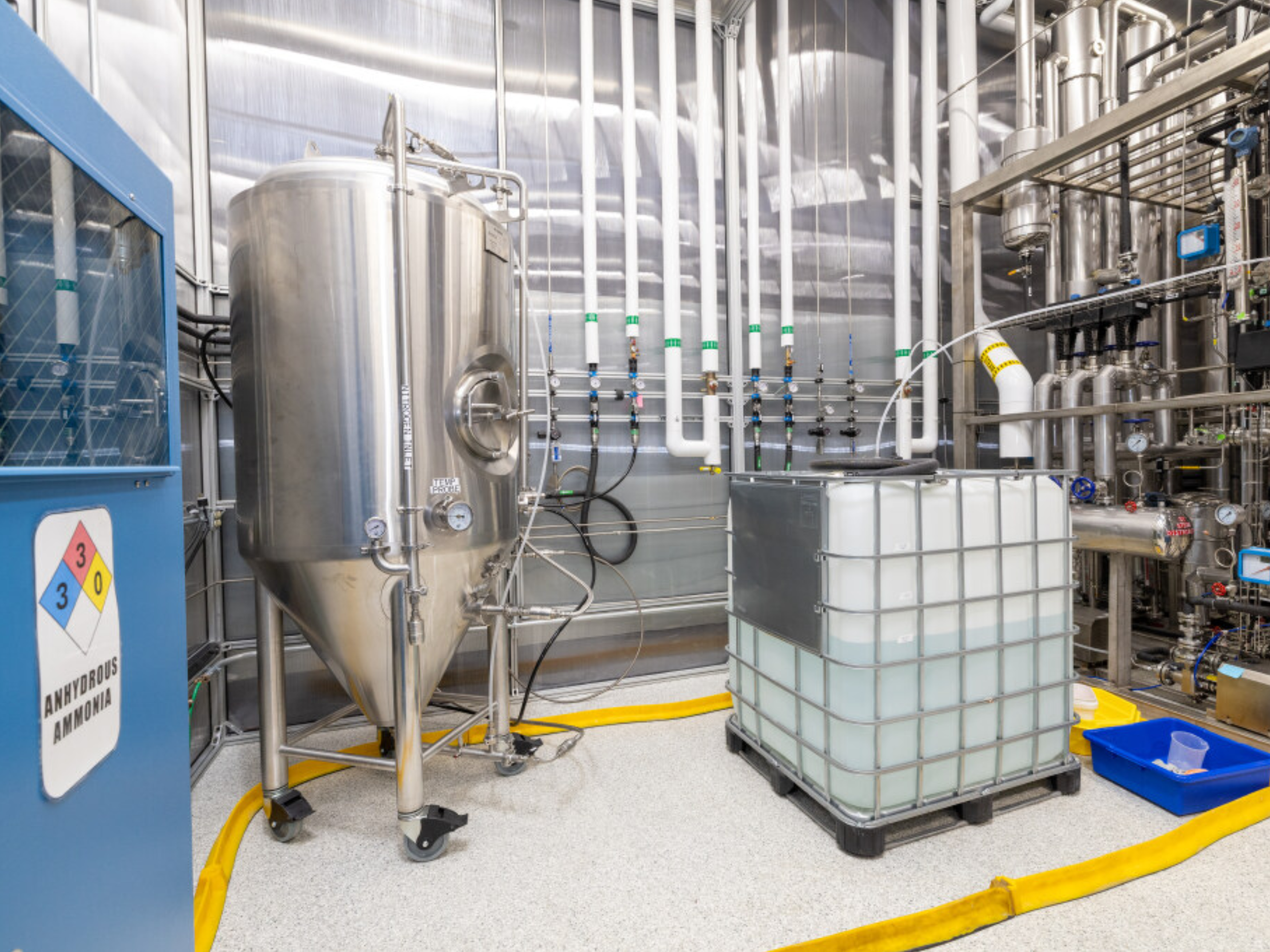 Industrial laboratory or manufacturing facility with metal tanks, pipes, gauges, a large white liquid container, and yellow hoses.