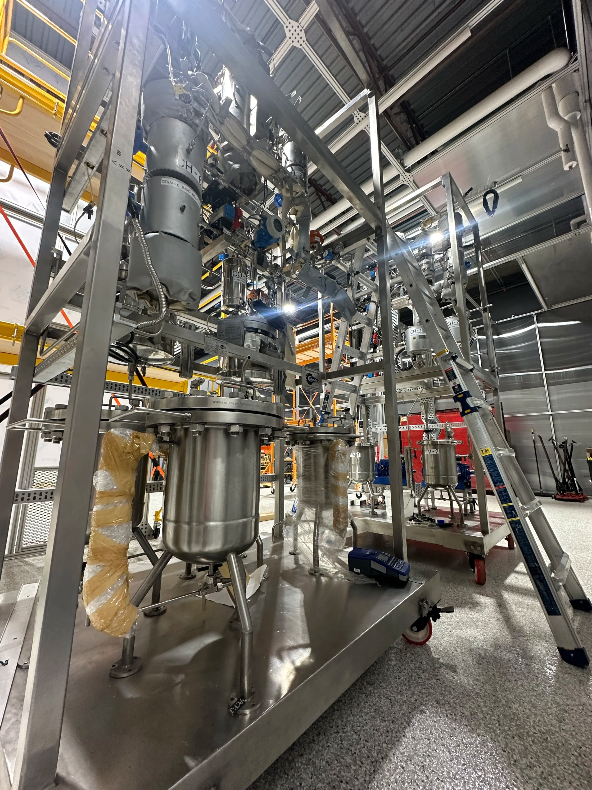 Industrial equipment with metal tanks, pipes, and a ladder inside a manufacturing or laboratory facility.
