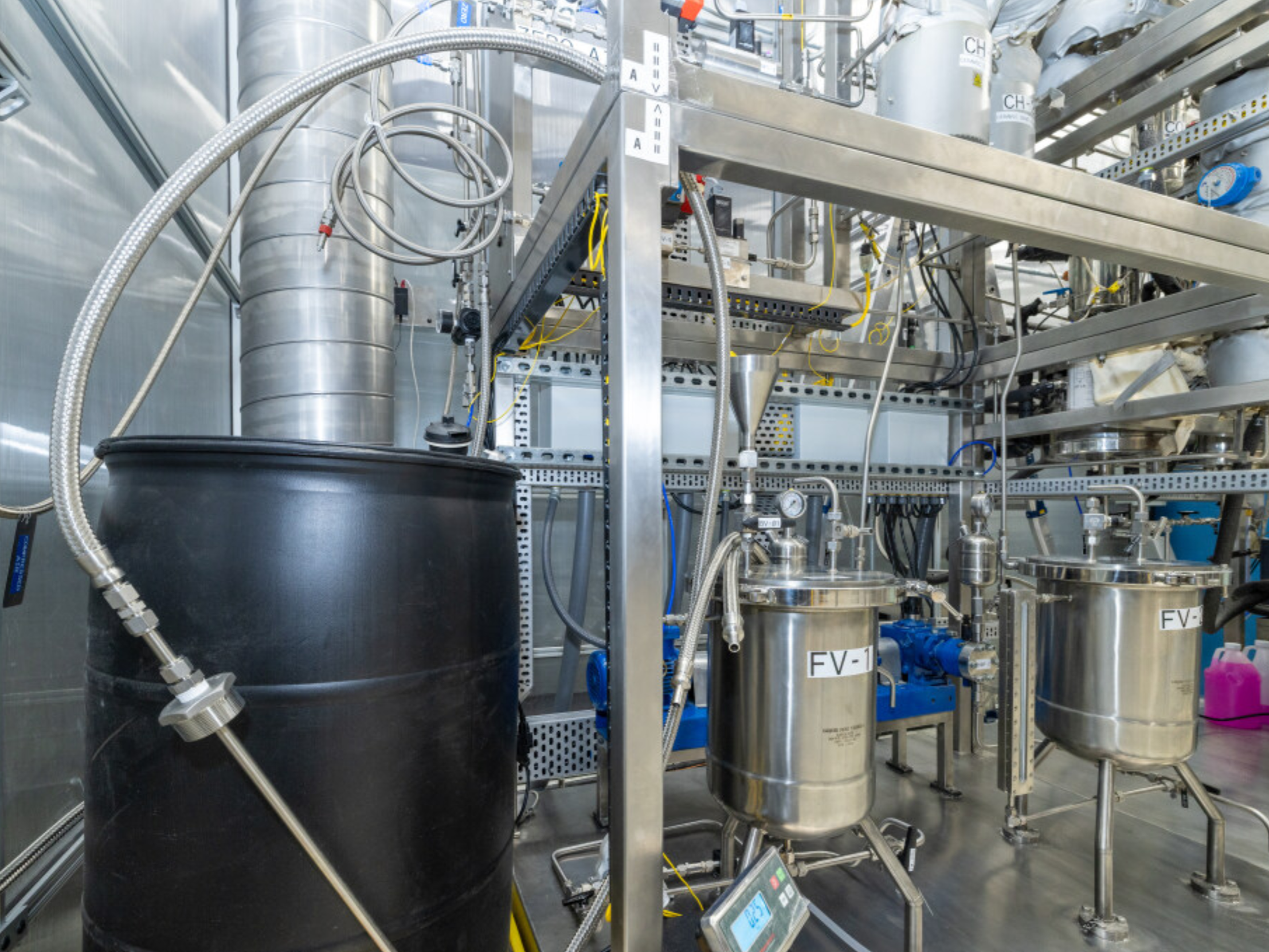 Industrial laboratory setup with metal containers, pipes, and wiring, including black and stainless steel vessels labeled FV-1 and FV-2.