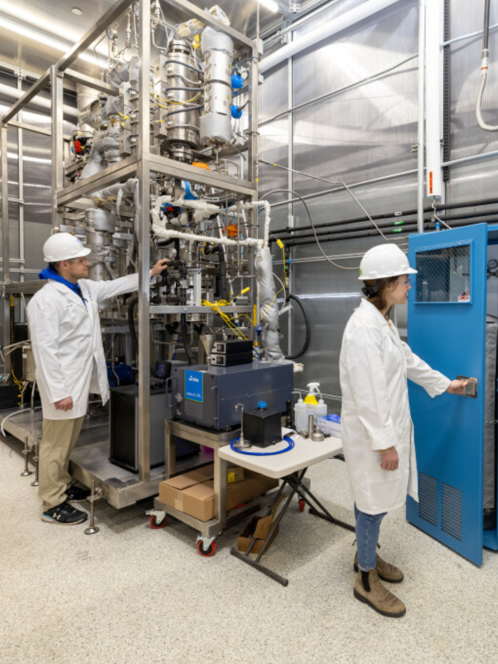 Two engineers in lab coats and helmets operating scientific equipment in a laboratory or industrial setting.