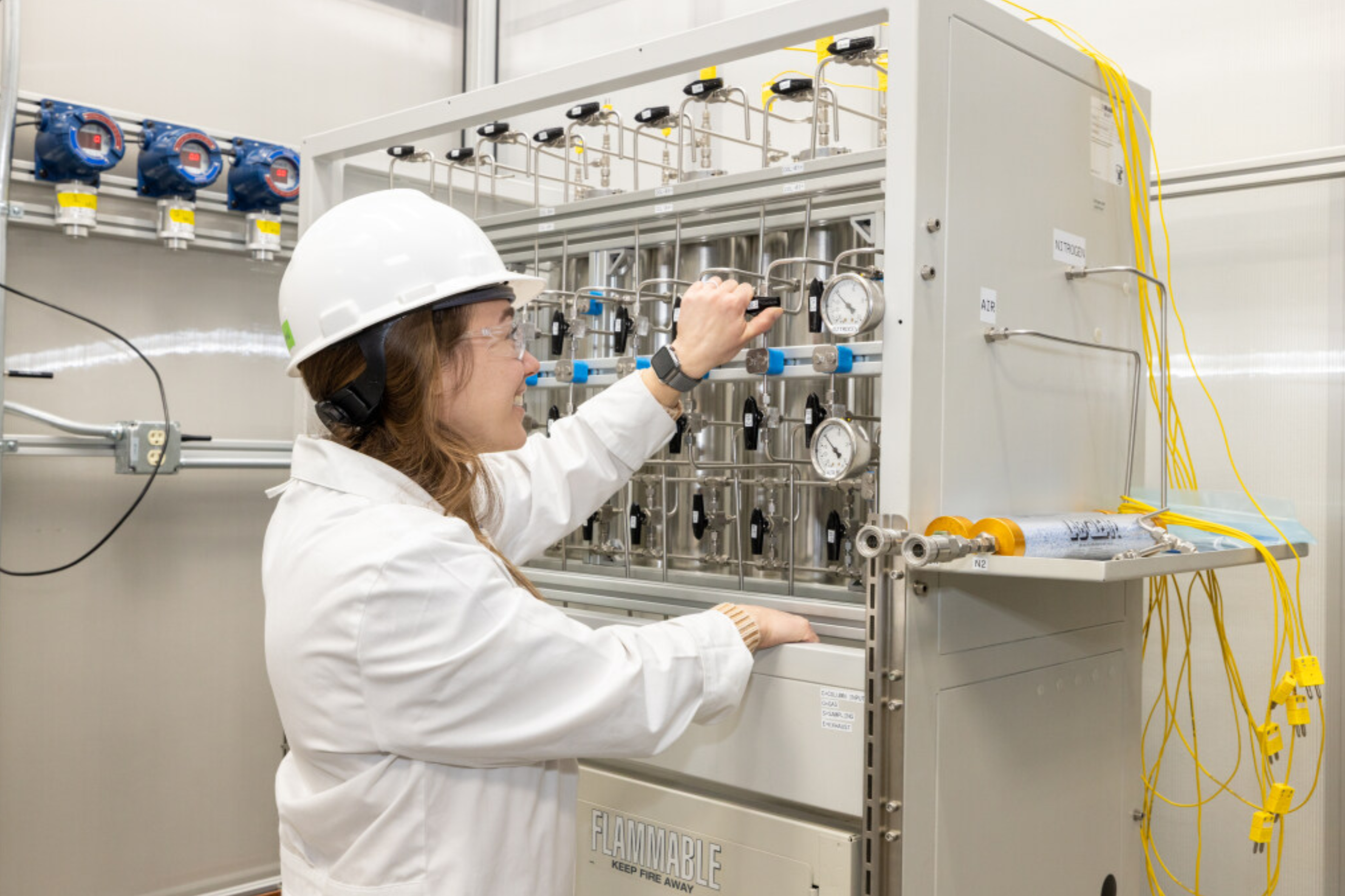A woman scientist wearing a white lab coat, safety helmet, and safety glasses adjusting pressure gauges on a complex industrial apparatus in a laboratory or manufacturing setting.