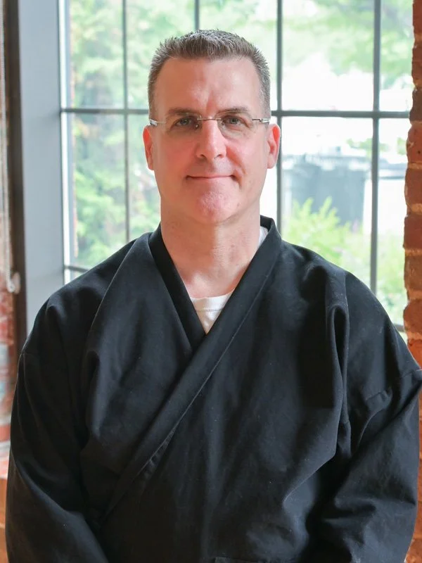 A man wearing a traditional black martial arts uniform, standing indoors near a large window with a blurred outdoor view.