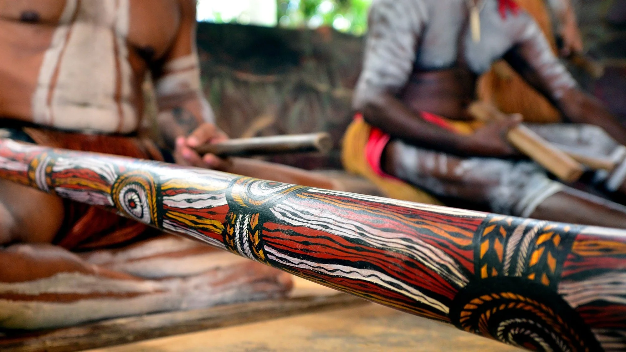 Close-up of a colorful, painted wooden spear or staff with intricate geometric and natural patterns, held horizontally. In the background, two individuals with dark skin and traditional tribal clothing sit outdoors, possibly engaged in a cultural activity or ceremony, in a natural setting with greenery.