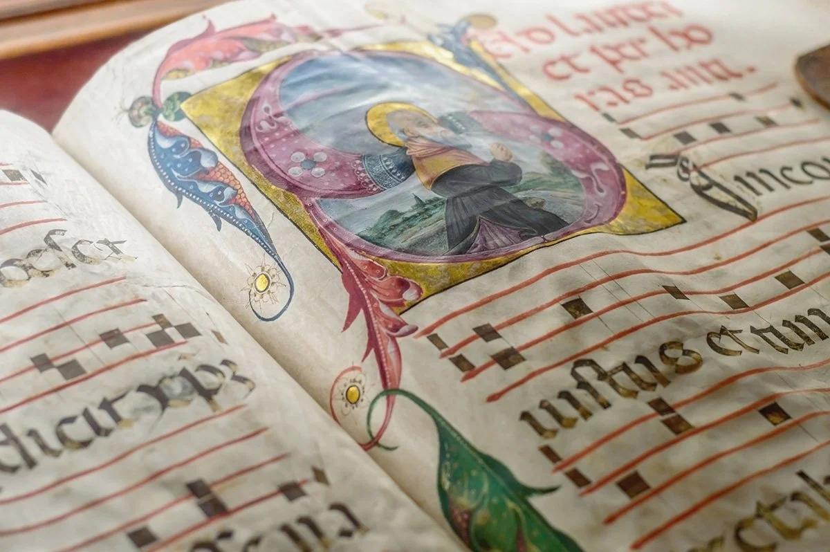 Colorful illuminated manuscript with musical notation and a detailed illustration of a religious figure, possibly a saint, inside a decorative frame.