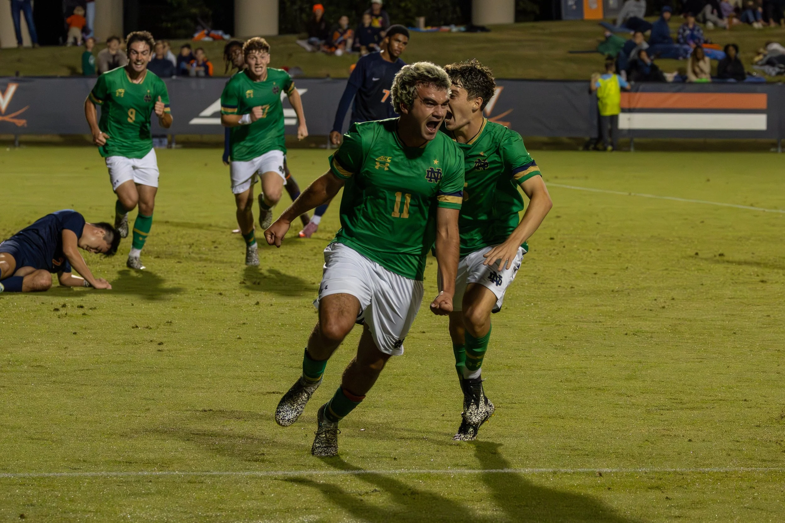 Soccer players celebrating on the field, with one player running and shouting in the foreground, teammates cheering behind him, and a fallen opponent on the ground.