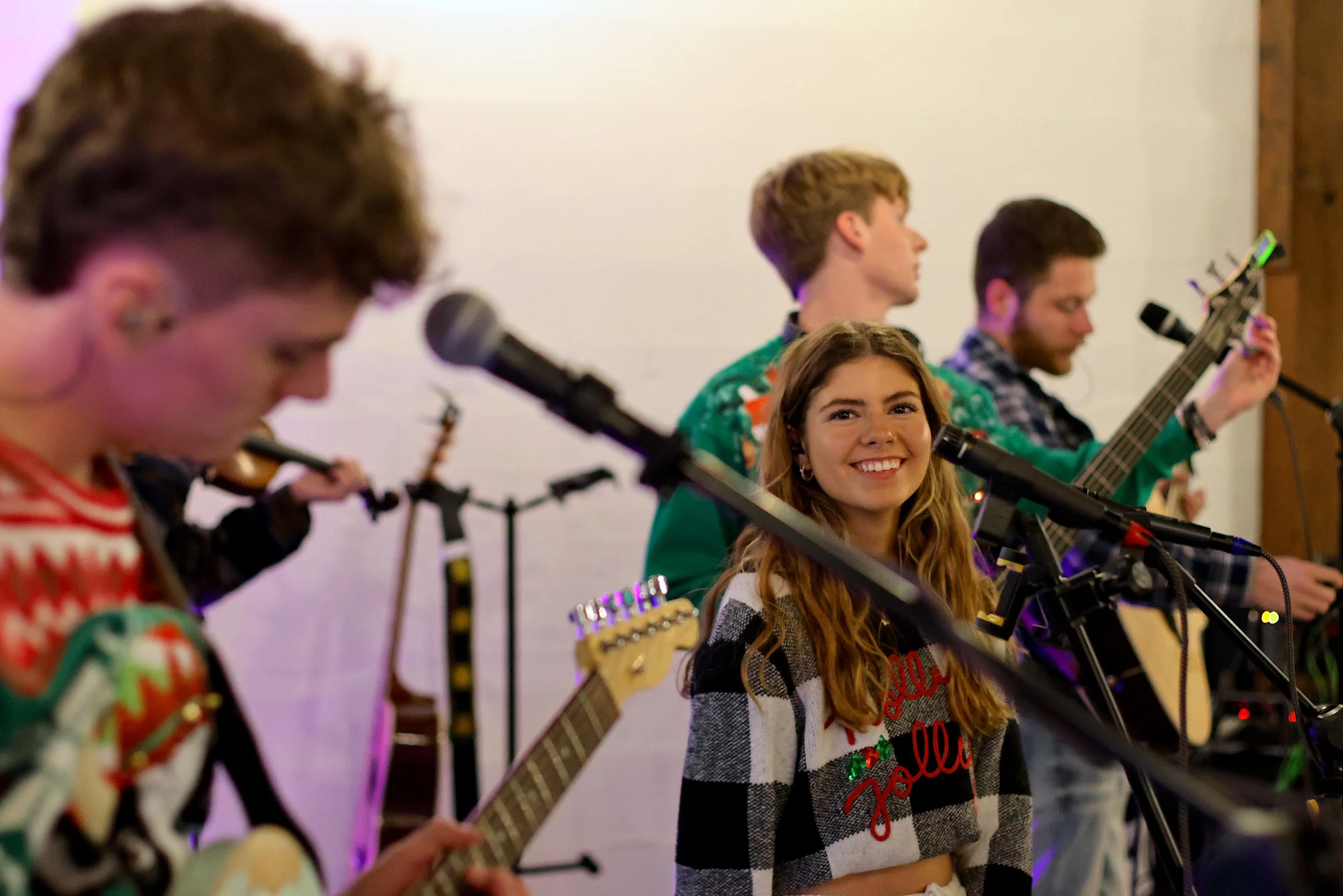 Young woman with long wavy hair smiling during a musical performance, surrounded by band members playing guitars and other instruments, in an indoor setting.