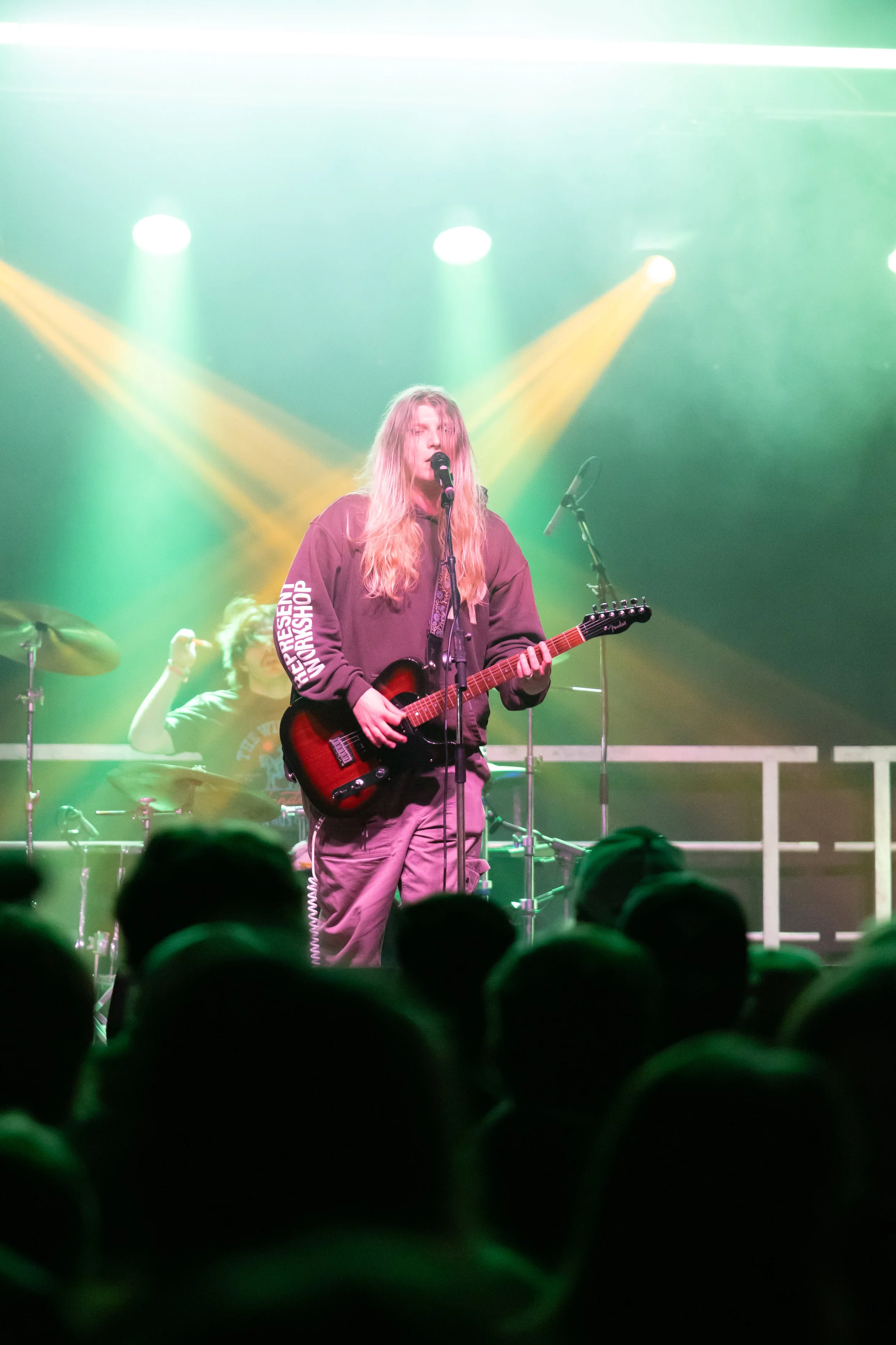 A female musician with long blonde hair singing and playing an electric guitar on stage, with a drummer in the background, colorful stage lights, and an audience in the foreground.