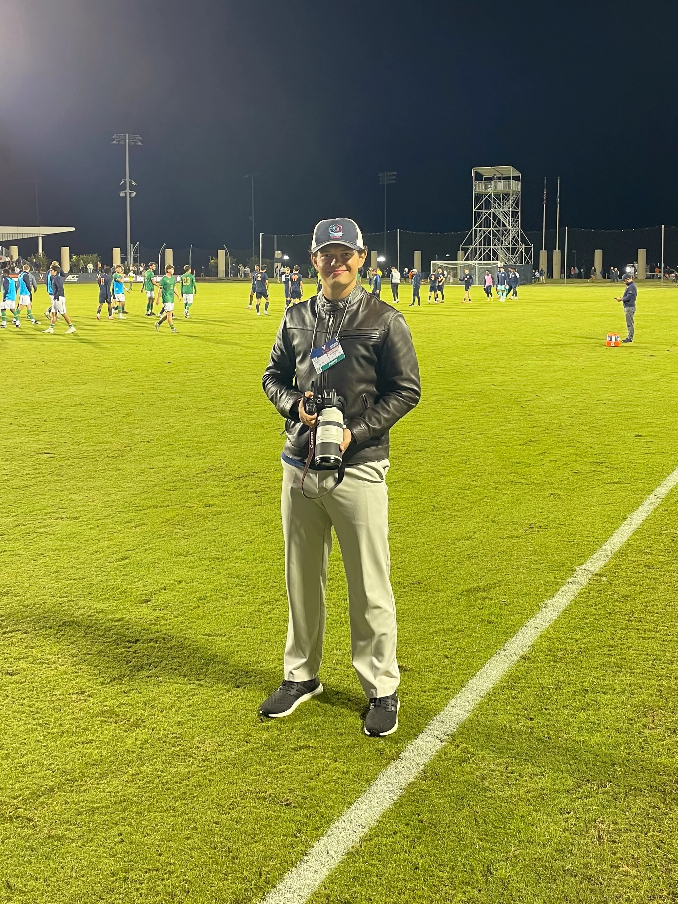 A photographer standing on a sports field at night, holding a camera with a large lens, with a team of football players warming up in the background.