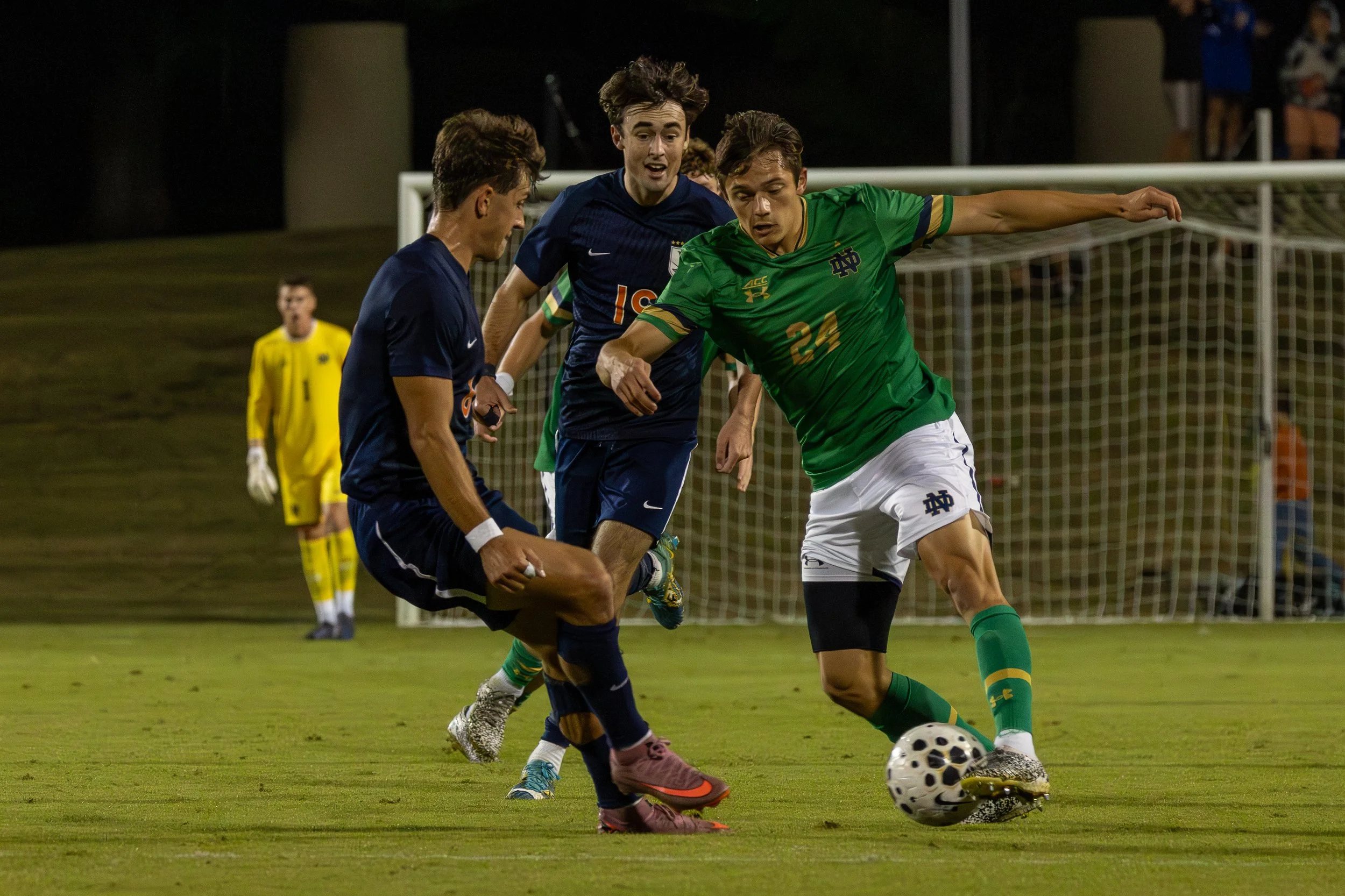 Soccer players battling for ball on field at night with goalkeeper in background.