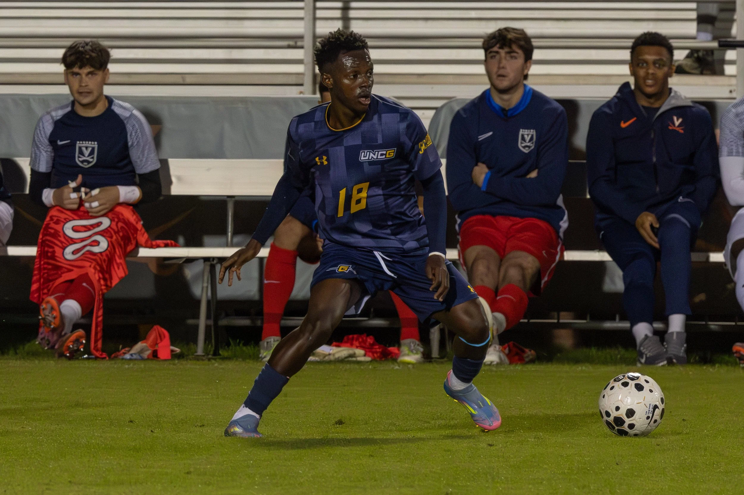 Soccer player in blue uniform with number 18 dribbling a soccer ball on field, with four seated players on bench in background, wearing athletic gear.