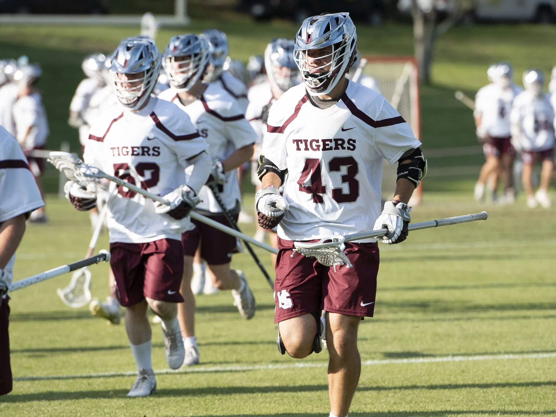 Lacrosse players from the Tigers team wearing white jerseys and maroon shorts, holding lacrosse sticks, running on a grassy field during practice or warm-up.