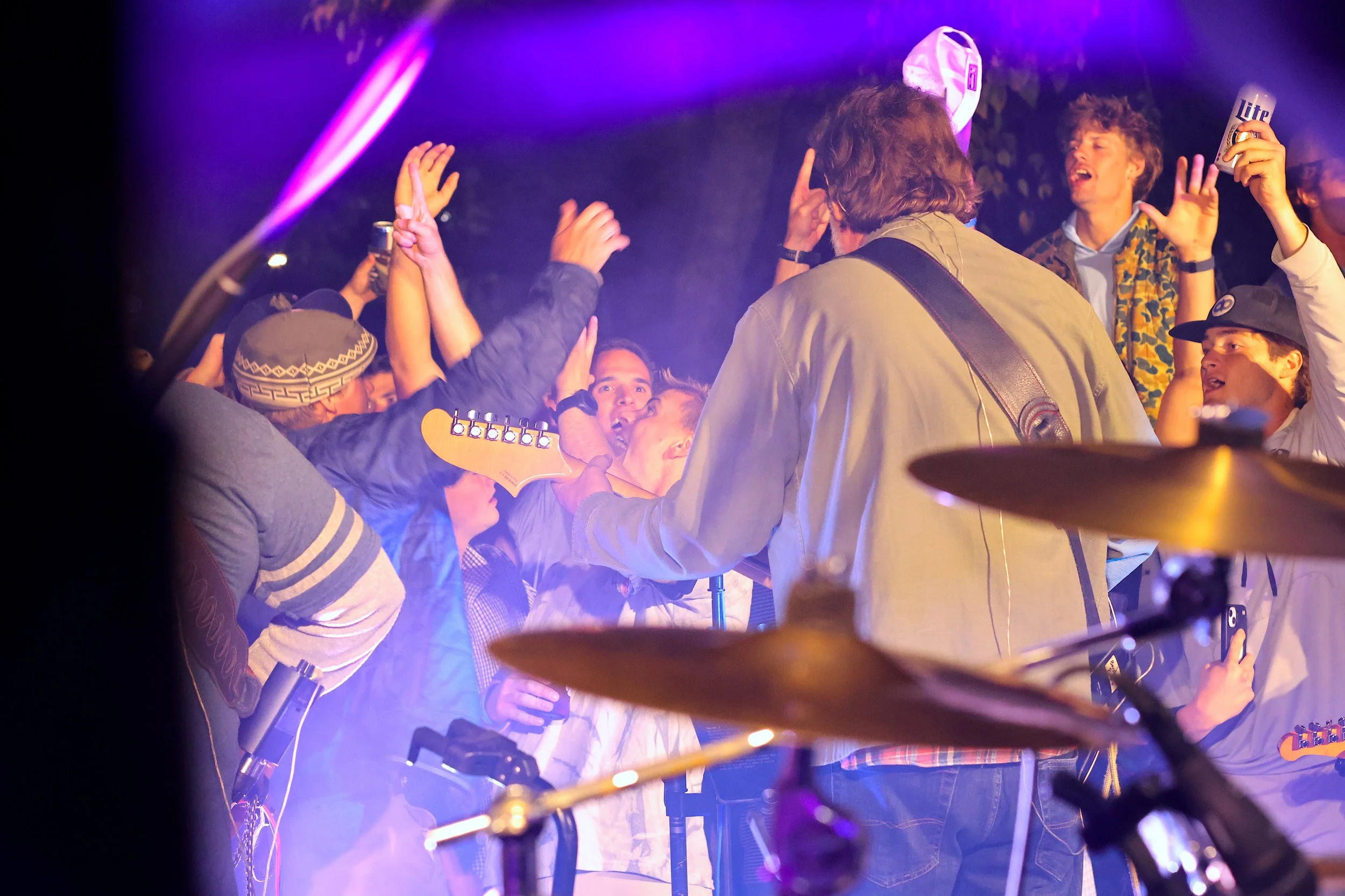 A lively concert scene with a band performing on stage, surrounded by an energetic crowd reaching towards the musicians. Purple and pink stage lights illuminate the scene, creating a vibrant atmosphere.