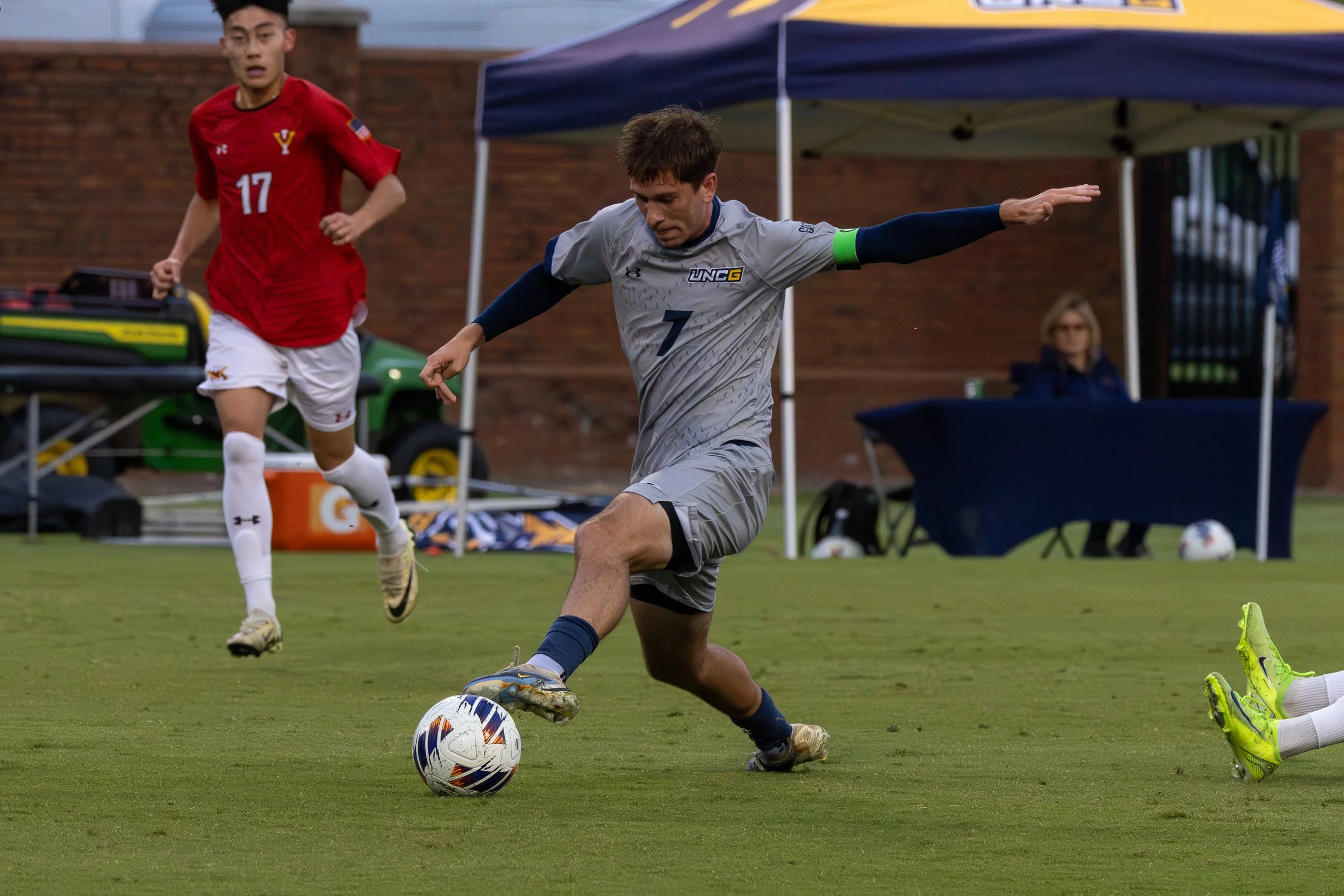 A soccer player in a gray uniform with number 7 kicking a soccer ball on the field, with another player in a red uniform running nearby and a woman seated under a blue tent in the background.
