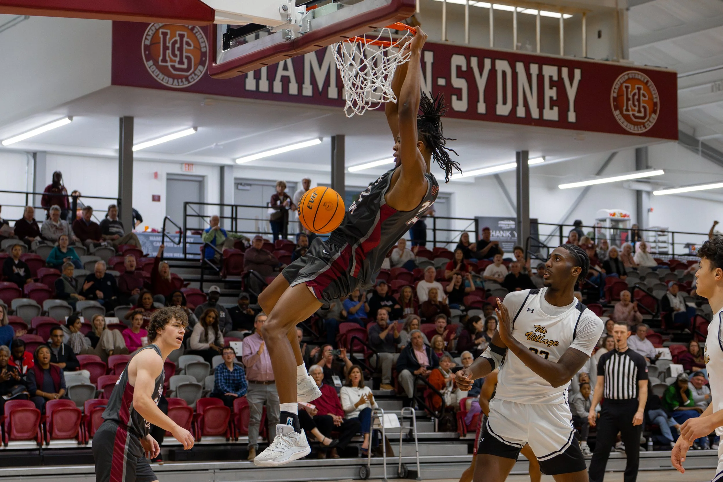 A female basketball player in a gray uniform is hanging from the basketball hoop after dunking the ball during a game. Two male players wearing white uniforms are nearby on the court, with one looking up at her and the other partially visible. The game is taking place in an indoor stadium with spectators seated in red and gray chairs.