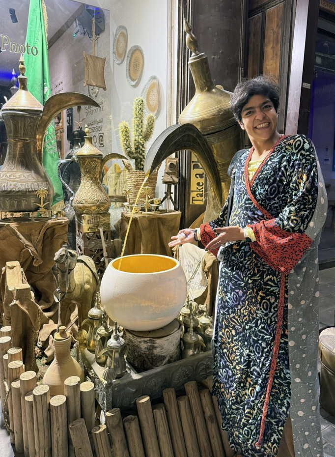 A woman in a colorful, patterned dress standing among various antique and decorative items, including brass vases, a ceramic bowl, and wooden objects in a shop display.